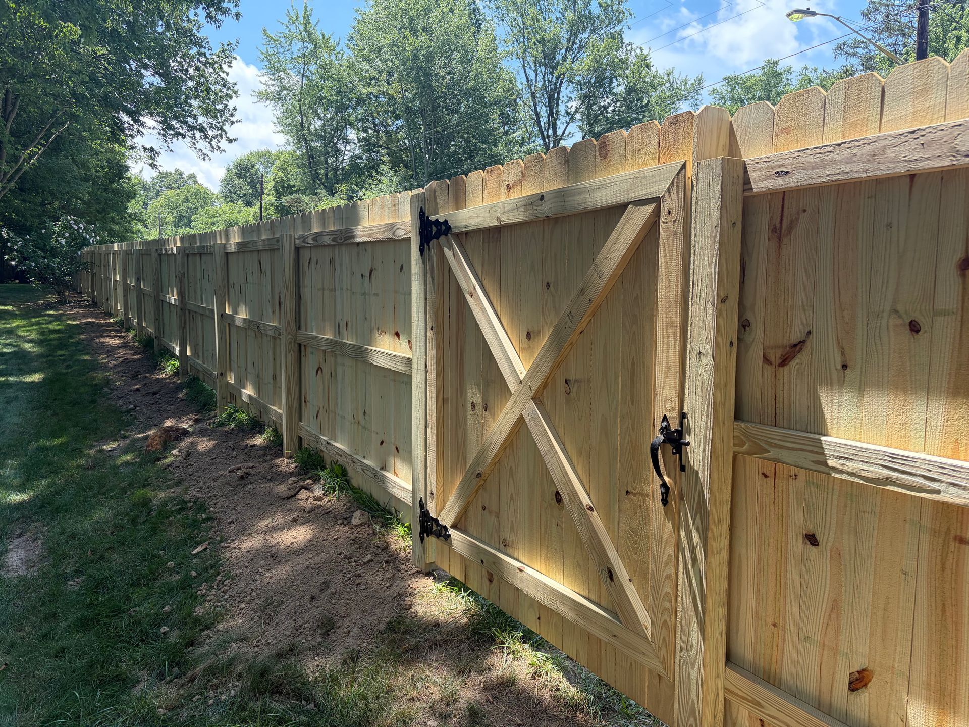 Wooden privacy fence with a large X-braced gate along a shaded dirt path.