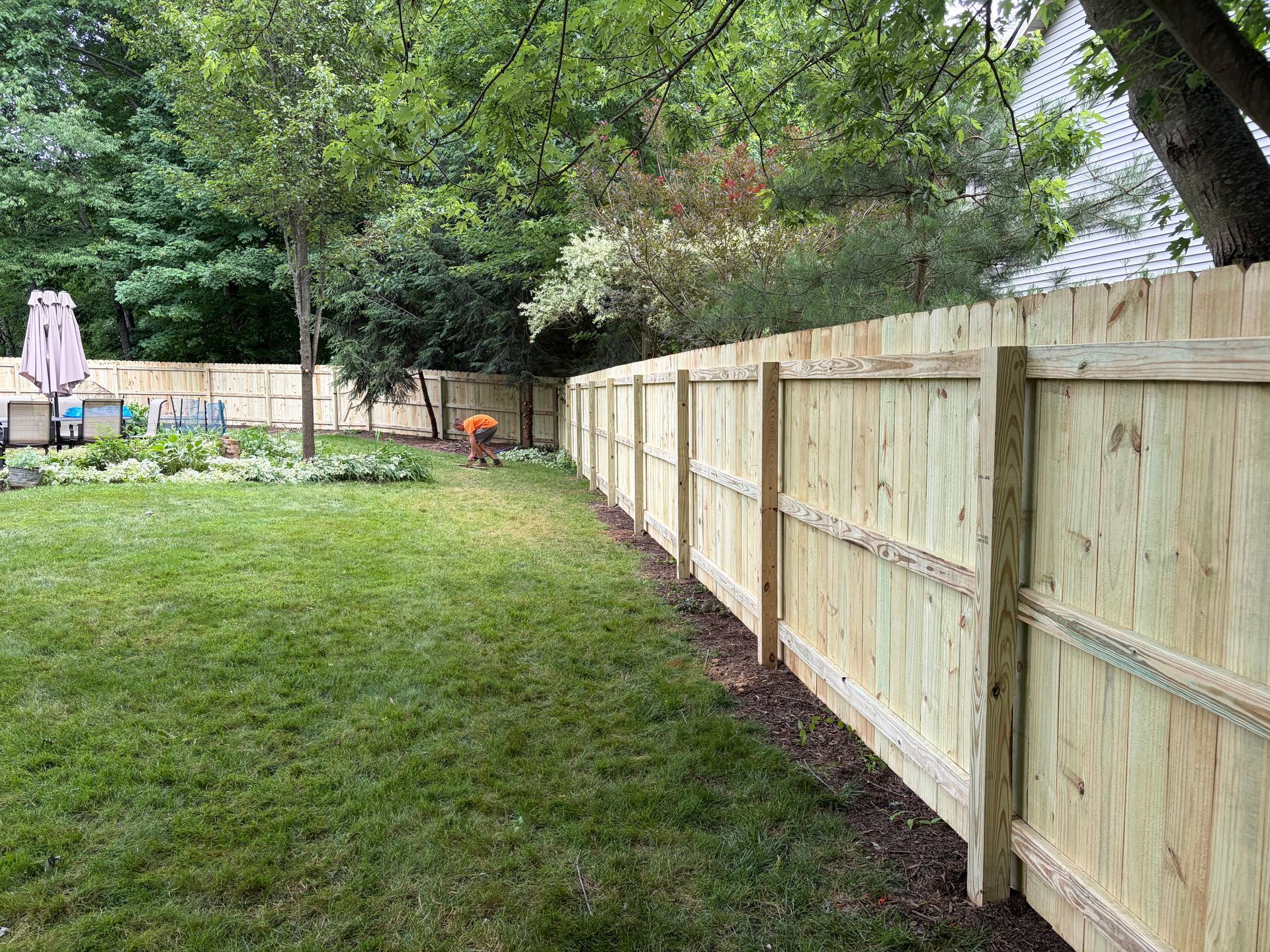 Backyard lawn beside a long wooden fence, with trees and garden beds in the background