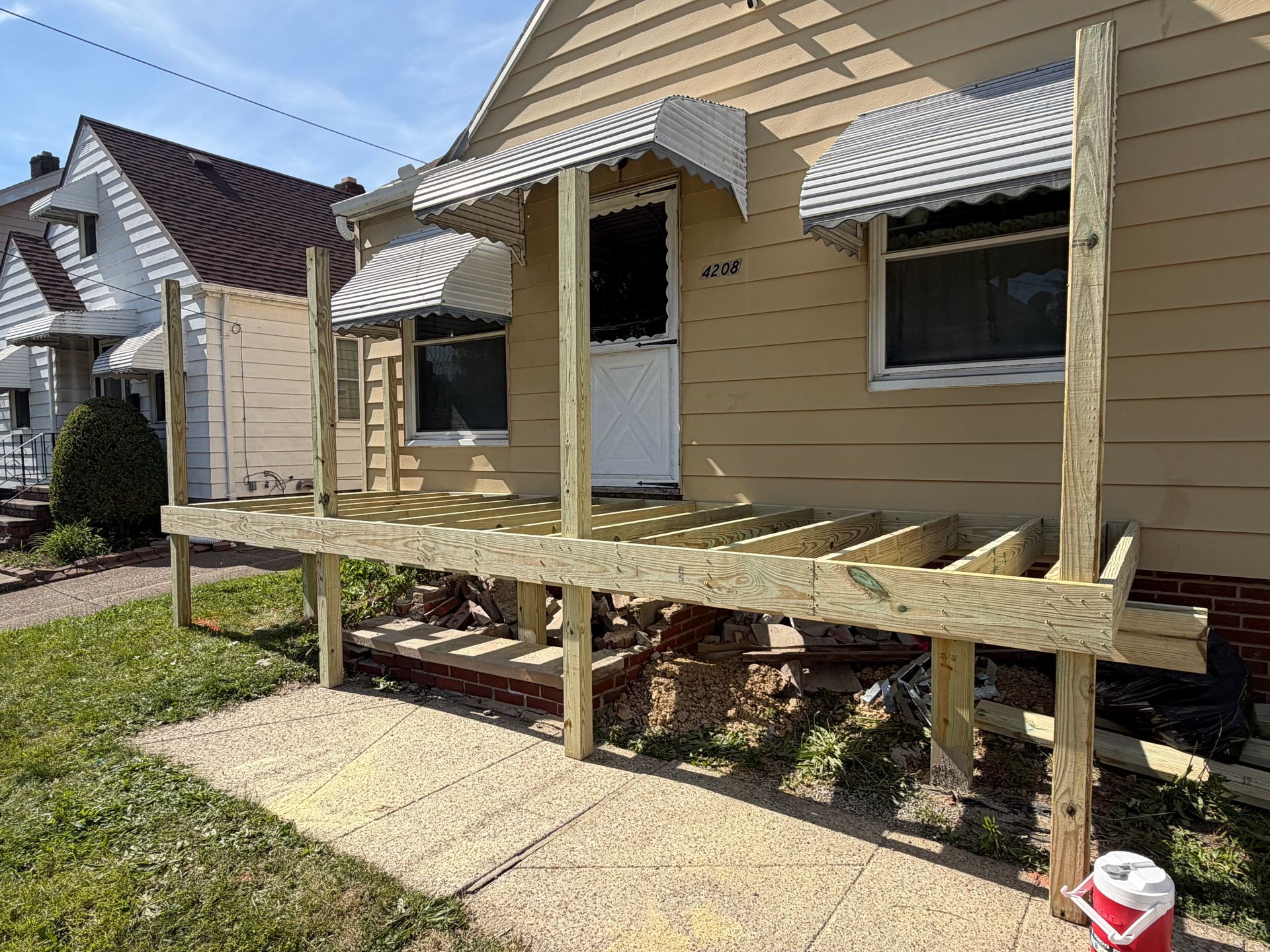Yellow house with a raised wooden porch under construction, beside a sidewalk and lawn