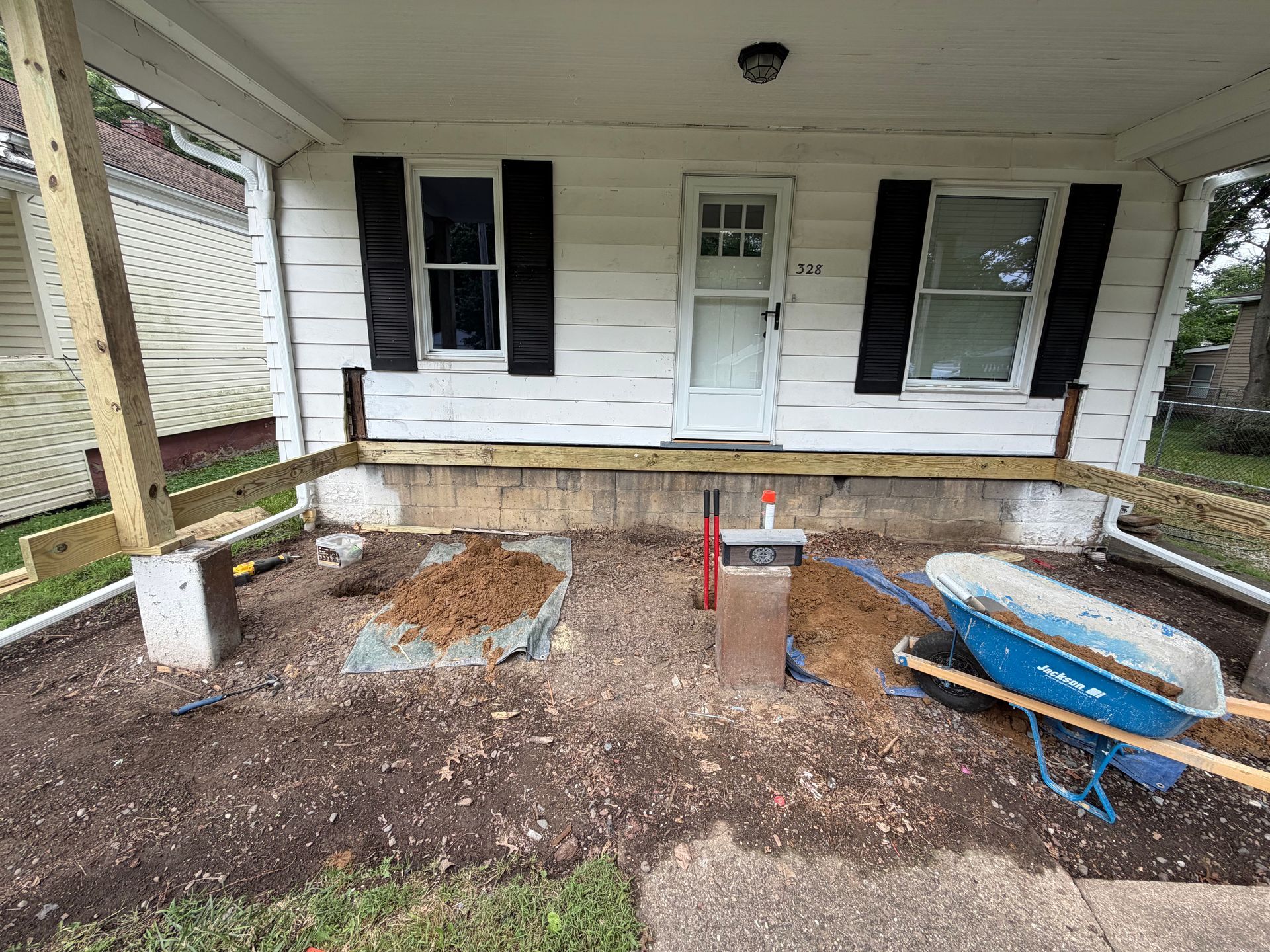 Front porch under renovation with exposed dirt, wheelbarrow, tools, and a white house facade.