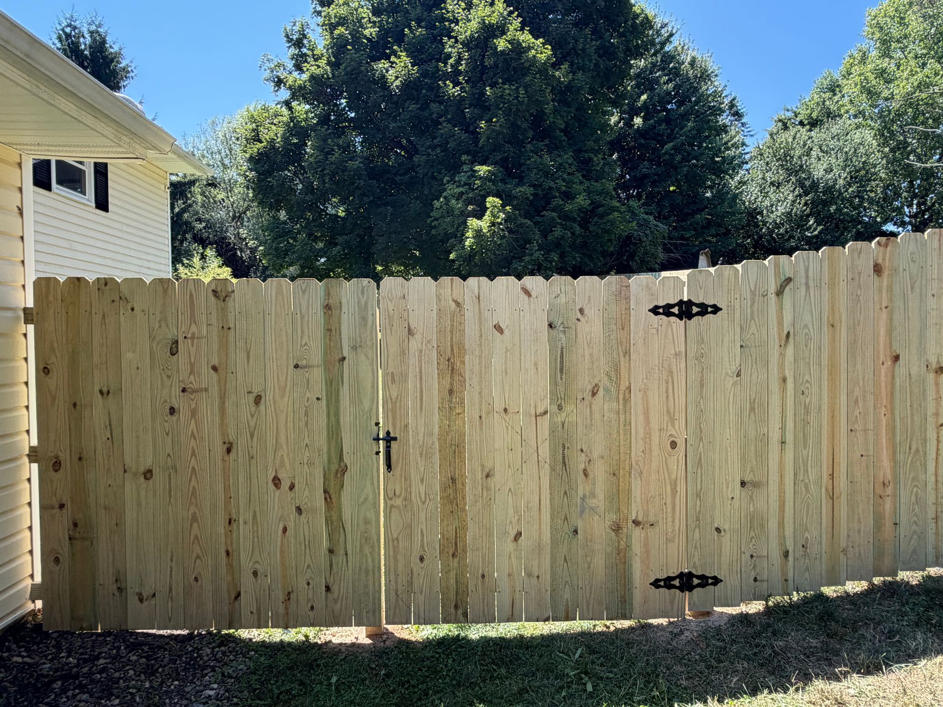Wooden backyard gate in a fenced yard beside a beige house, with trees and blue sky behind it