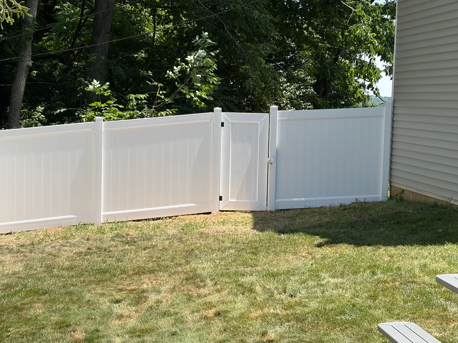 White vinyl fence with a gate in a grassy backyard beside a house and trees