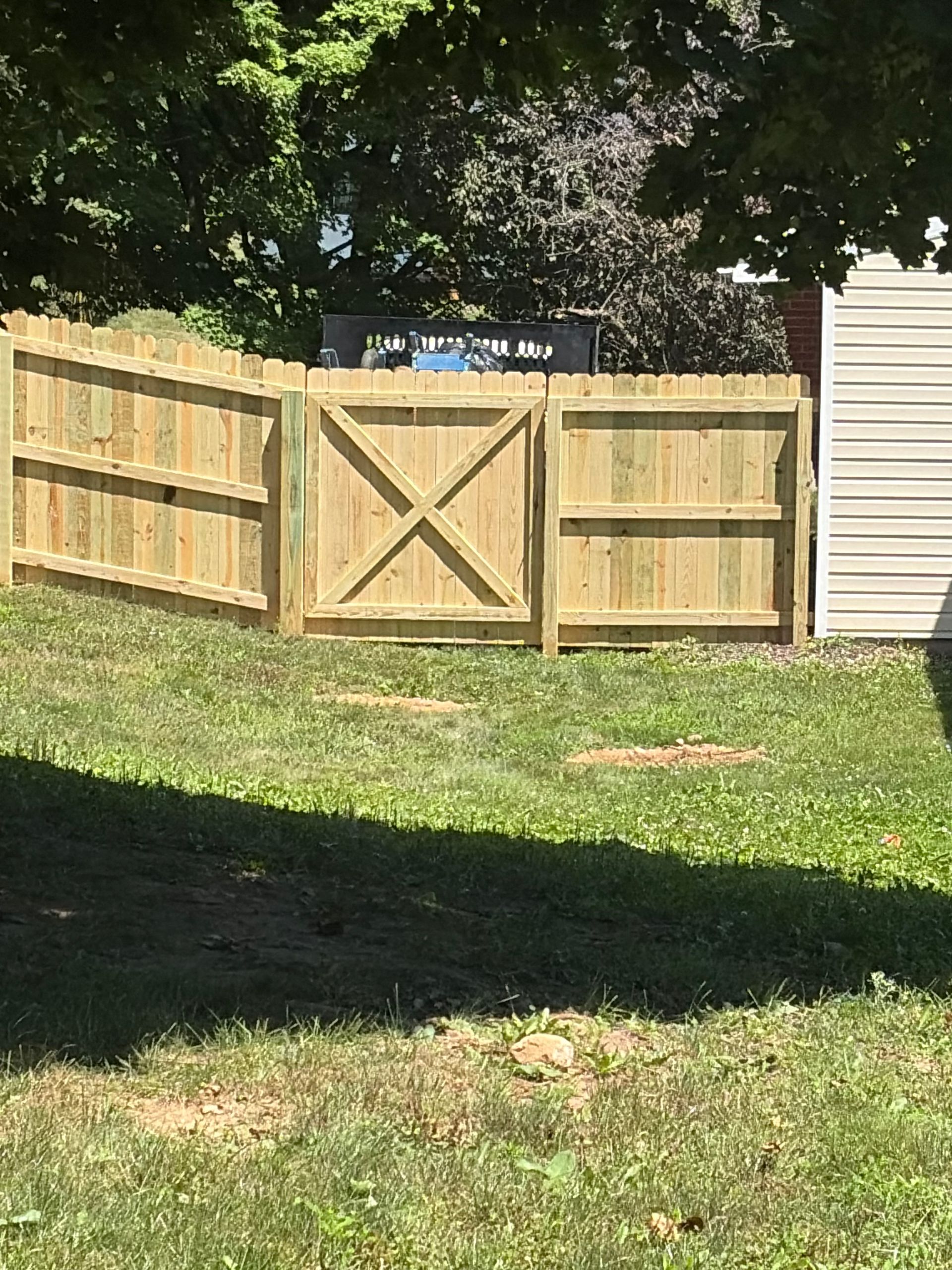 Wooden backyard fence with a centered gate, sunlit grass, and trees casting shade