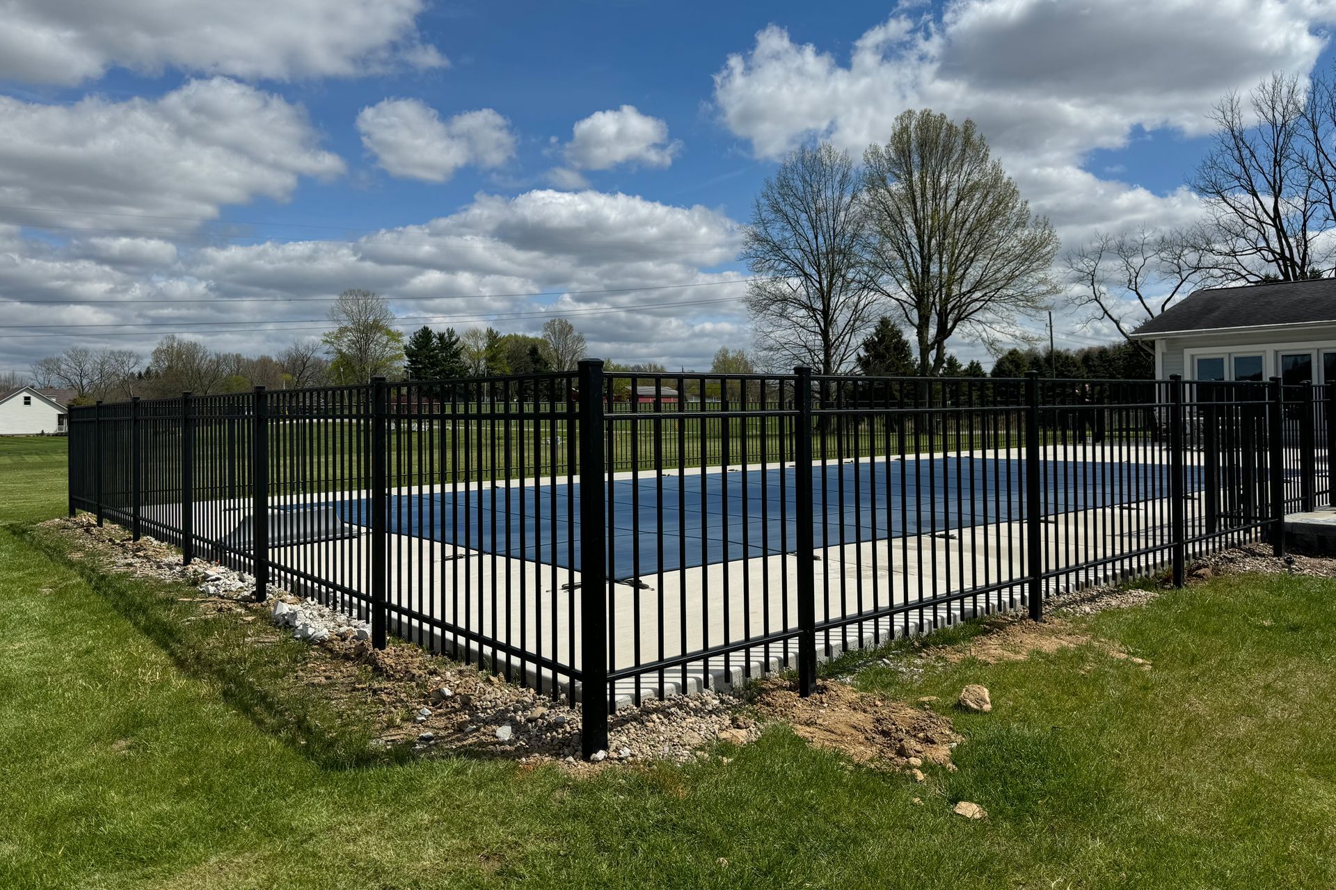 Black metal fence surrounding a blue-covered pool in a grassy yard on a sunny day.