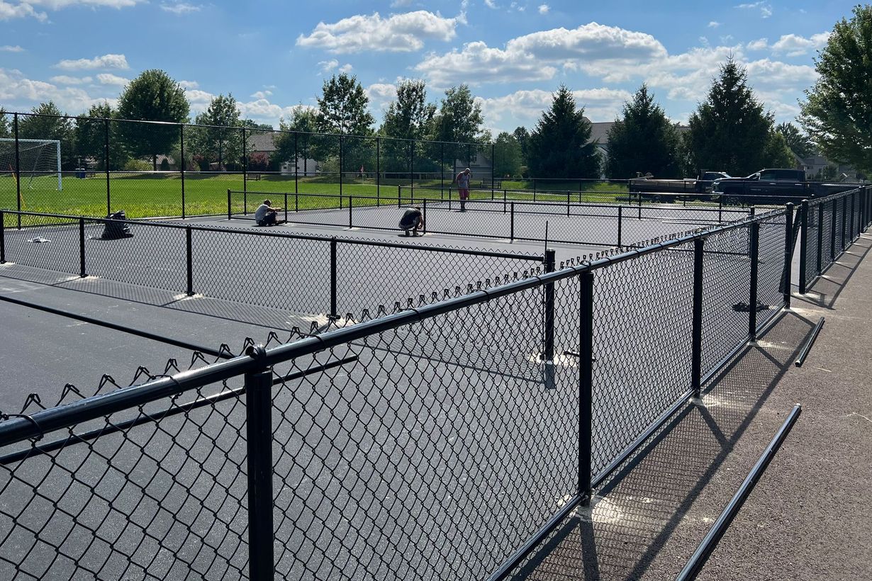 Black chain-link fence surrounds a dog park. Trees and houses are in the background on a sunny day.