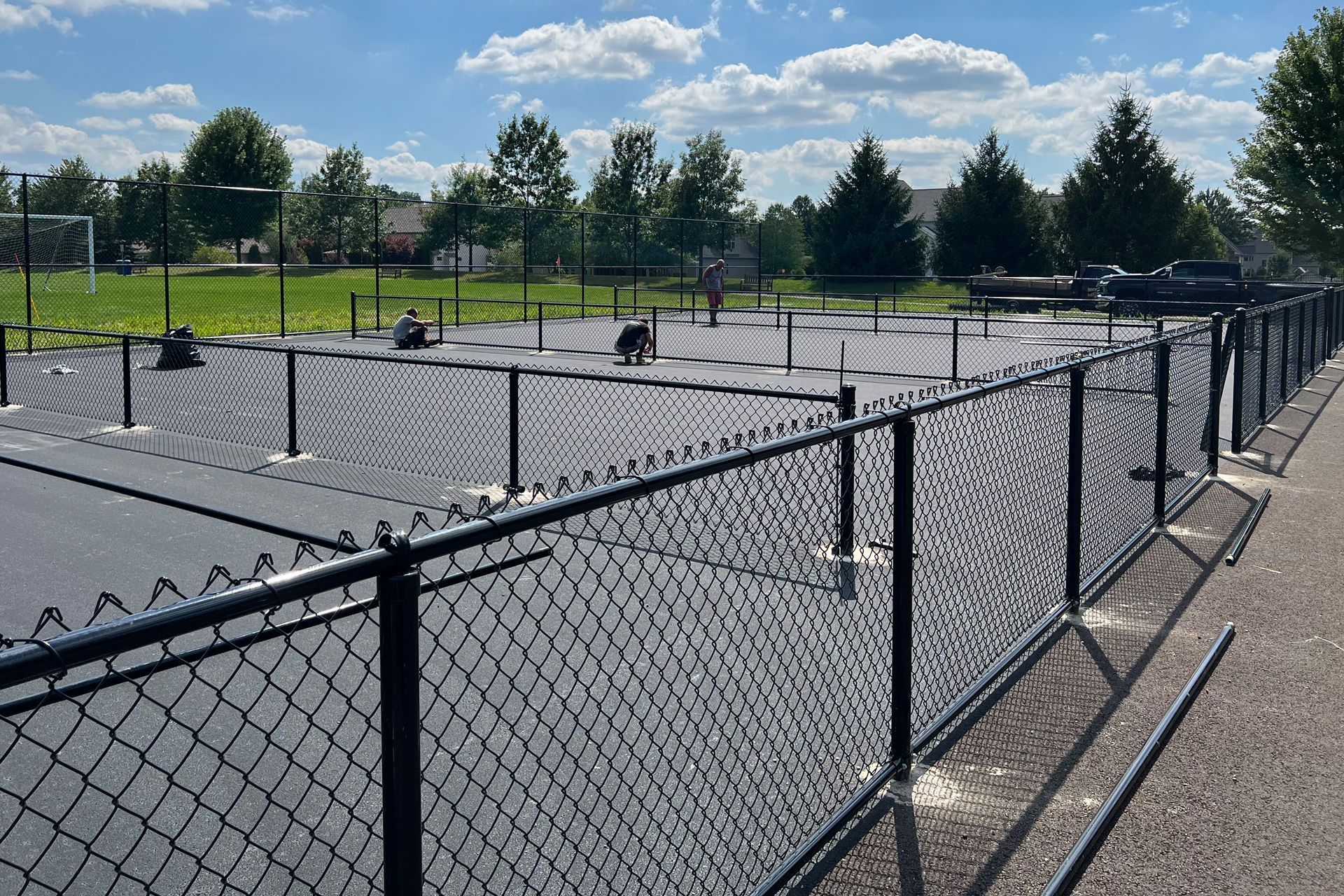 Black chain-link fence surrounds a dog park. Trees and houses are in the background on a sunny day.