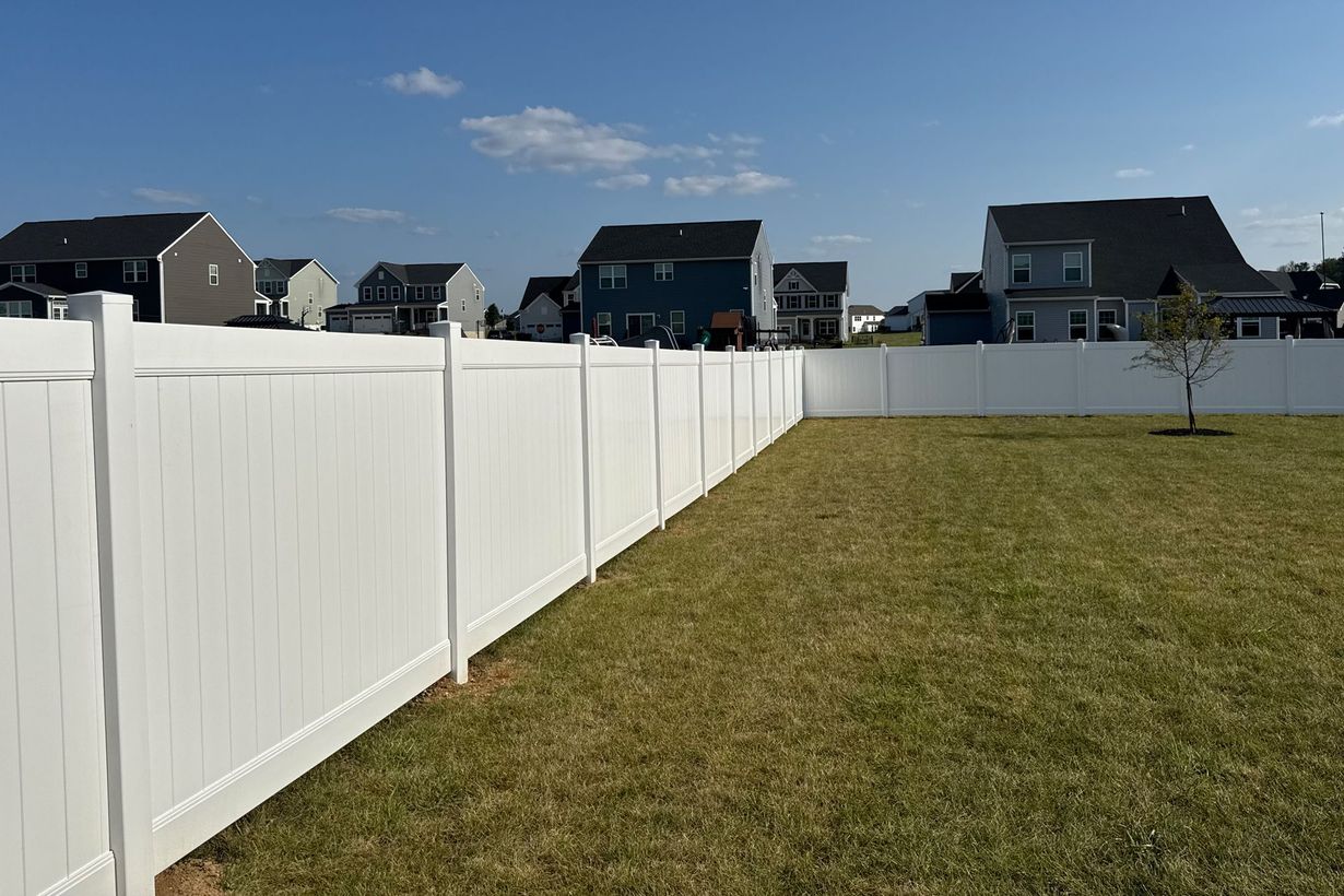 White fence encloses a grassy backyard, with houses in the background under a blue sky.