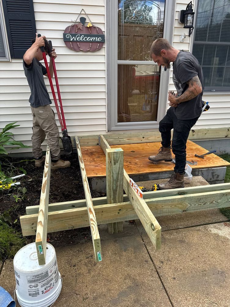 Two men building a wooden ramp at a house entrance. One operates a jackhammer, the other examines the construction.