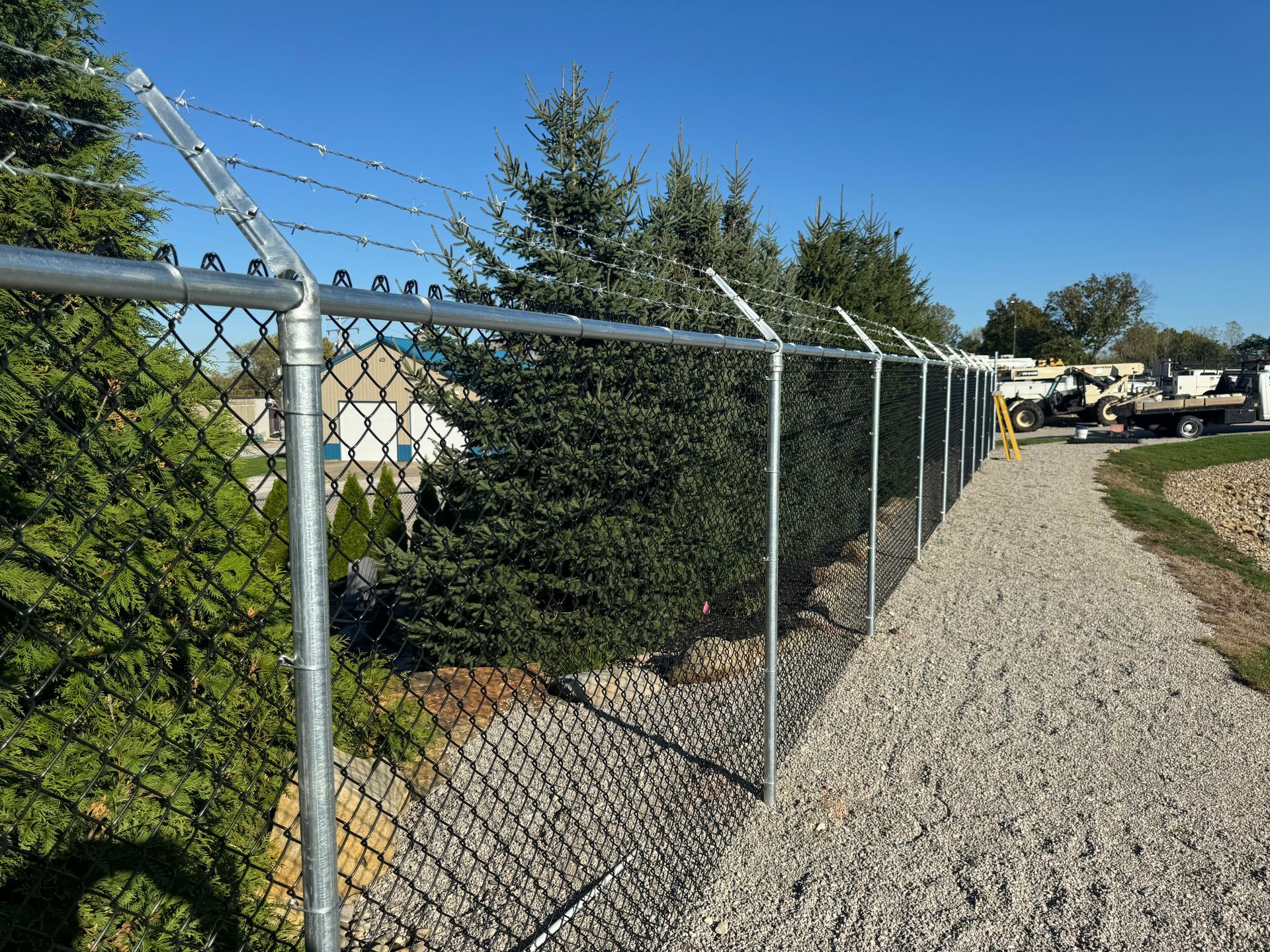 Chain-link fence topped with barbed wire alongside a gravel path, trees and a clear blue sky.