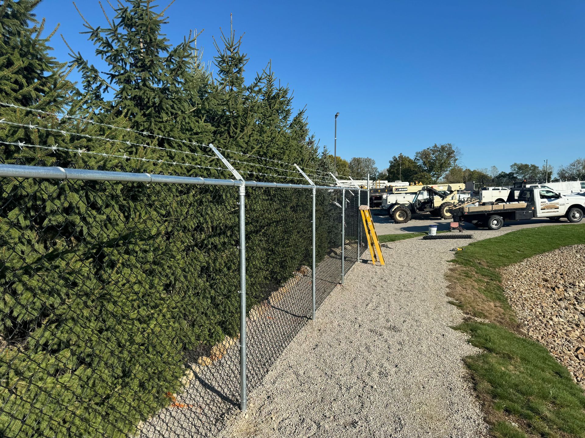 Chain link fence with barbed wire atop, bordering a gravel path and dense green hedge. Vehicles in the background.