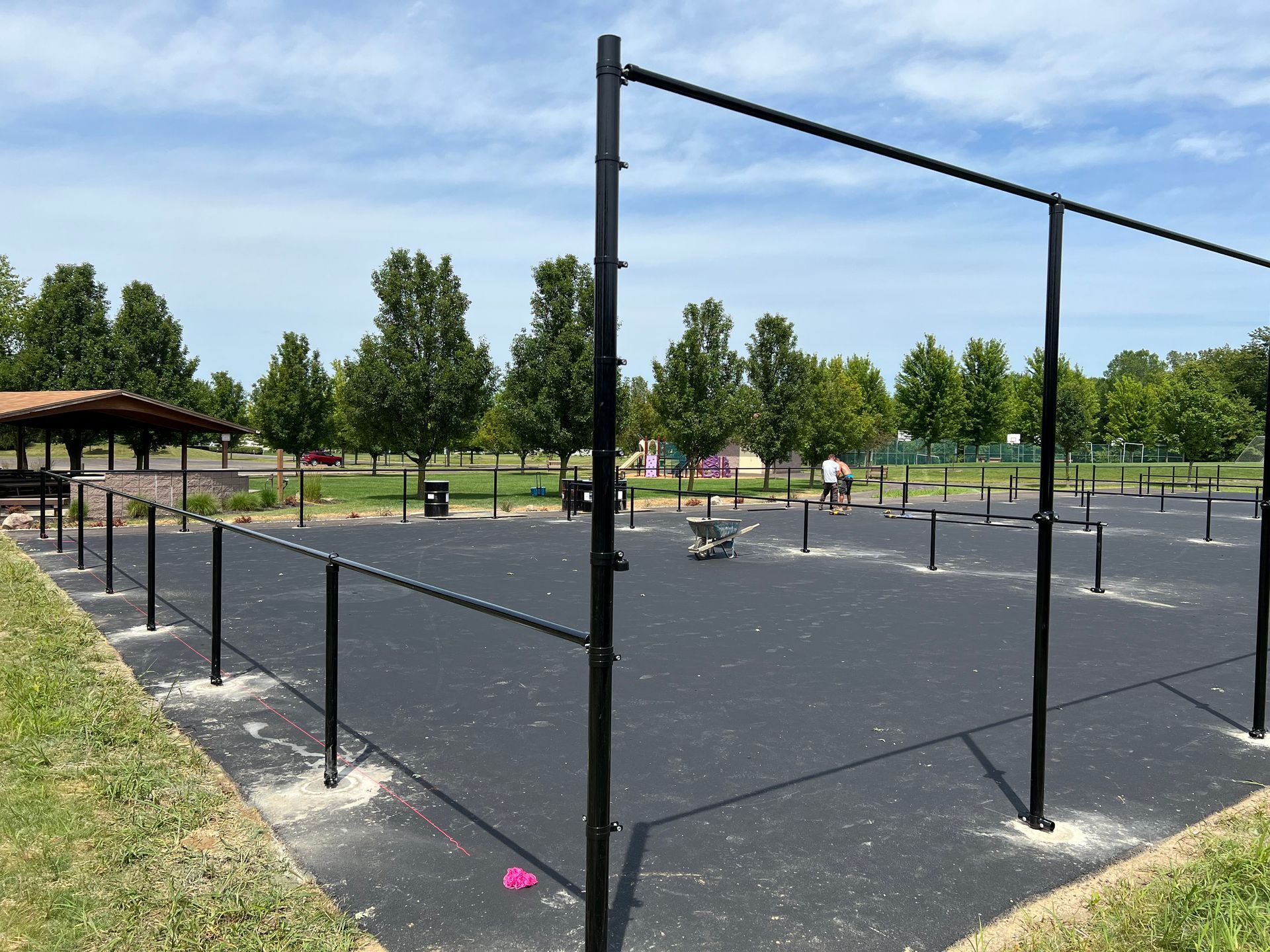 Black fenced area with exercise equipment at a park, trees and pavilion in background.