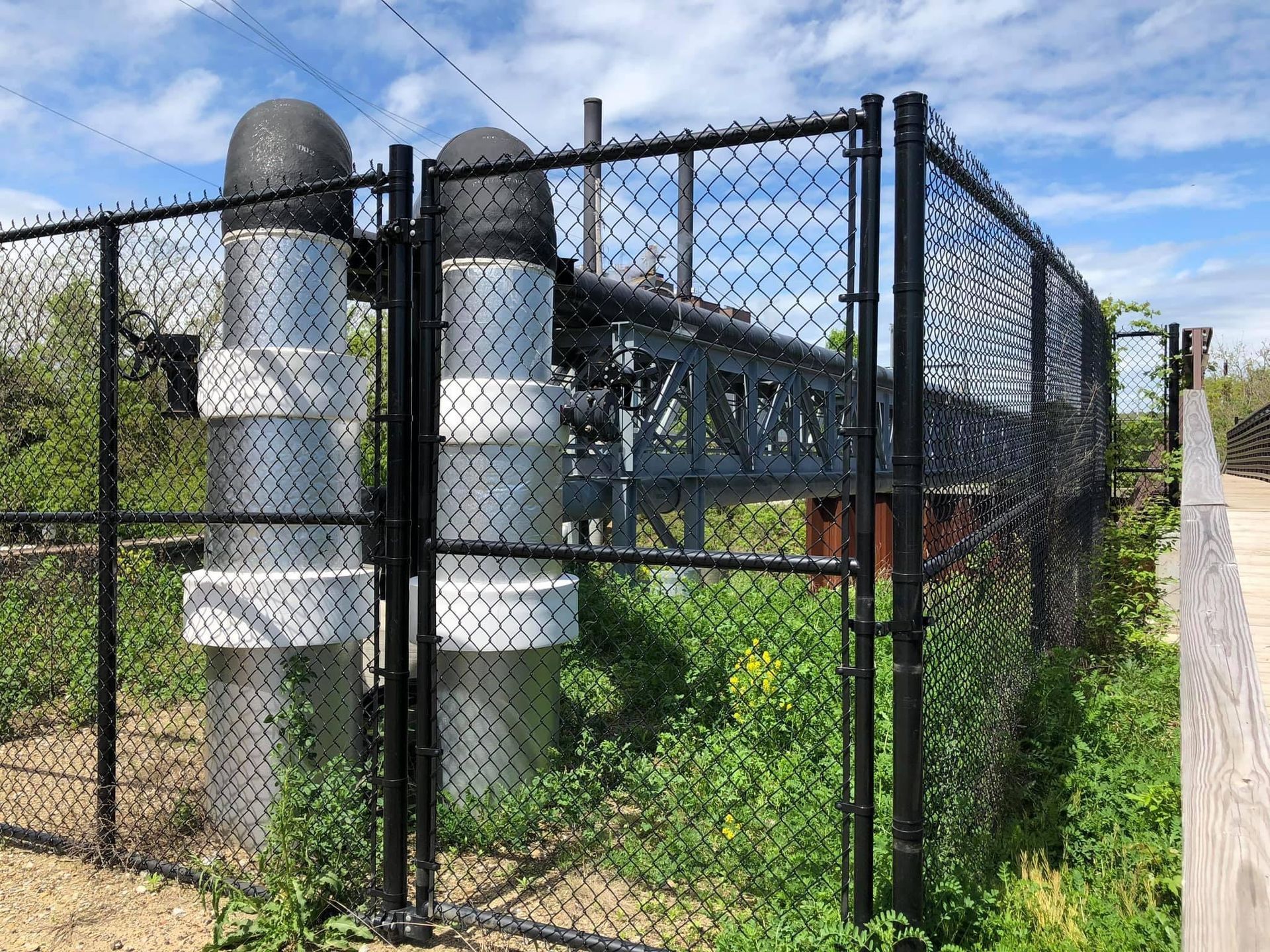 Black fence surrounding tall, silver pipes with black tops and a metal structure in the distance, with green foliage and a blue sky.