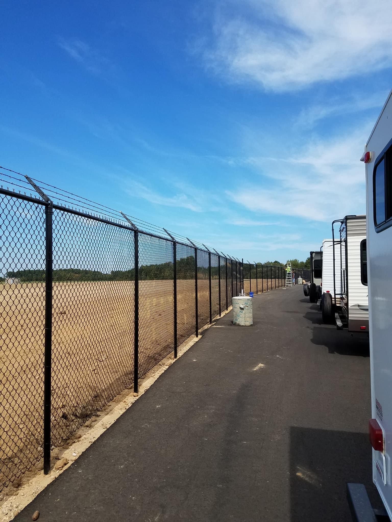 Black fence with barbed wire, RVs parked on right, blue sky.