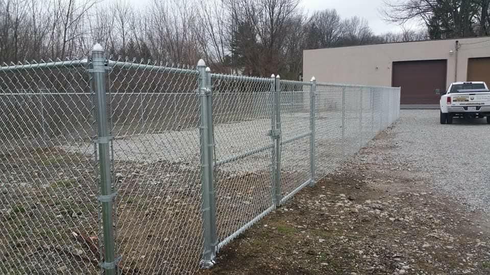 Chain-link fence surrounding a gravel lot with a building and truck in the background. Cloudy day.