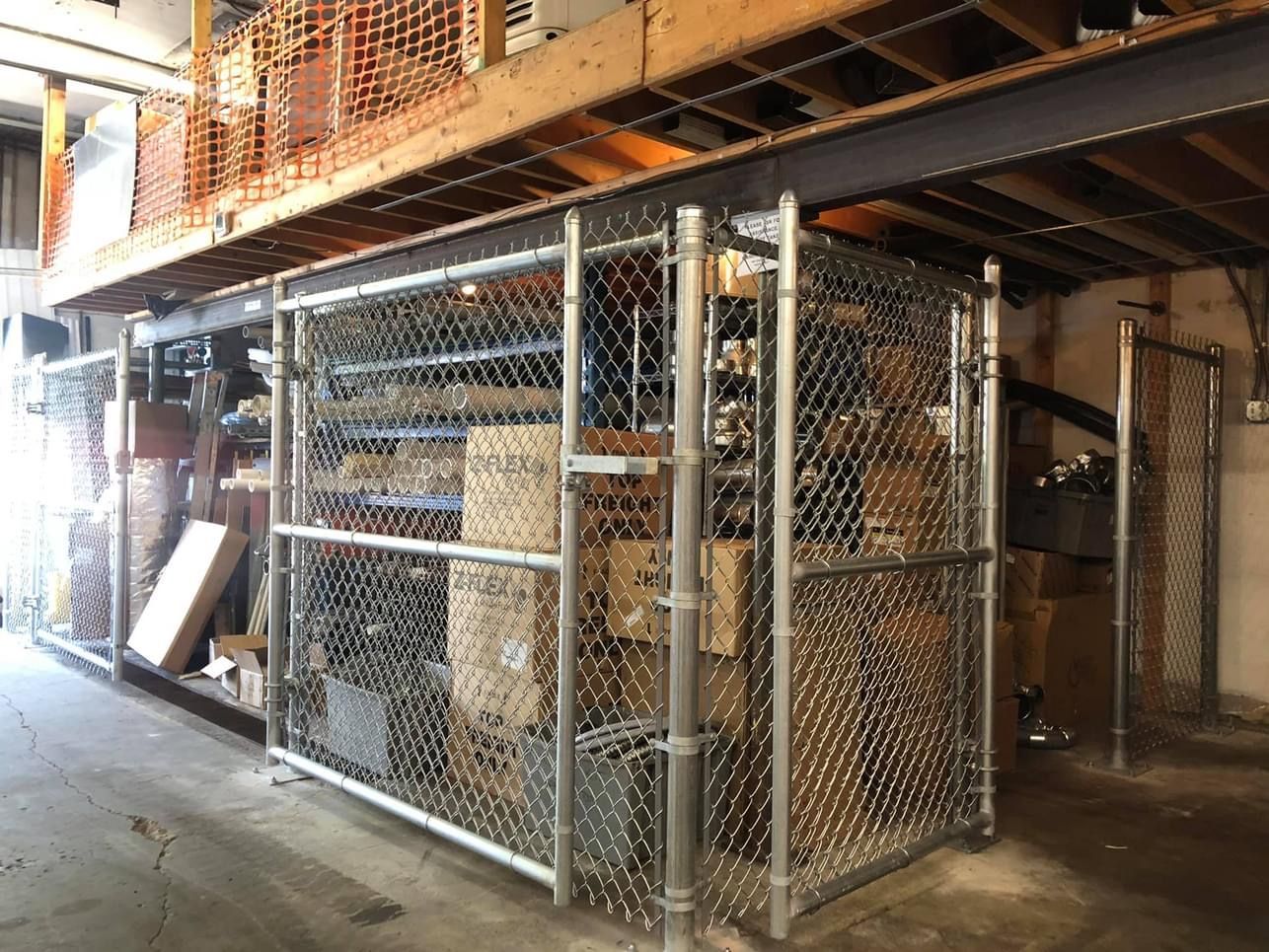 Chain-link cages hold boxes and items under a wooden loft. Industrial setting, gray metal and wood tones.