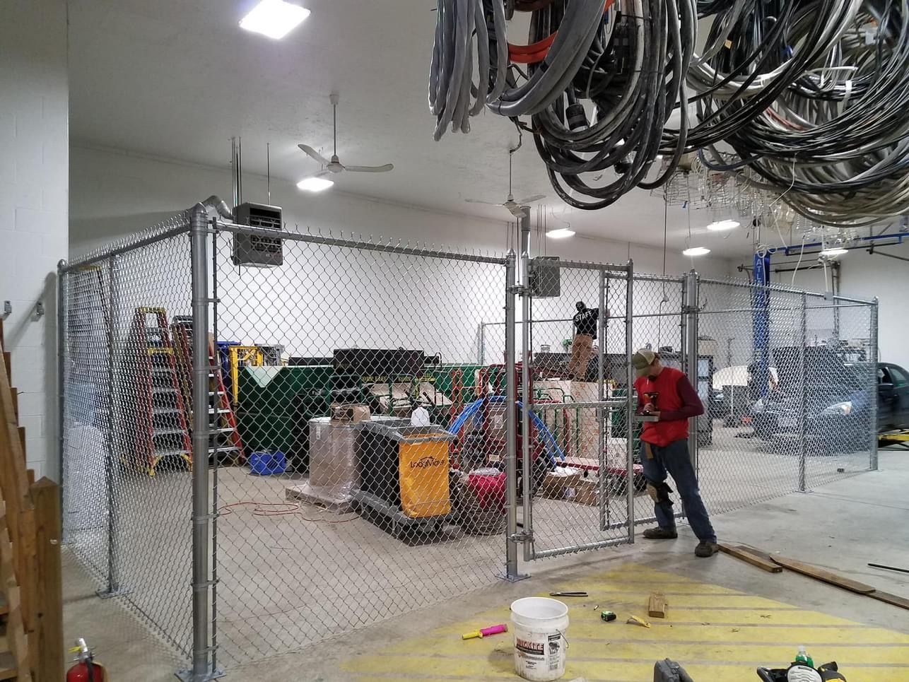 A man in a red shirt opening a chain-link gate inside a warehouse with equipment and supplies.