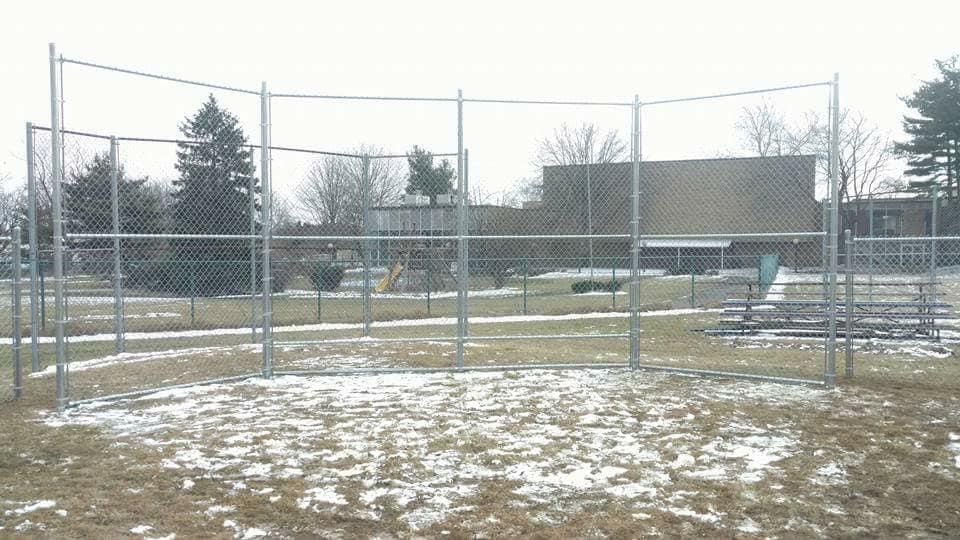 Chain-link fence surrounding a snow-covered area, trees and buildings in background.