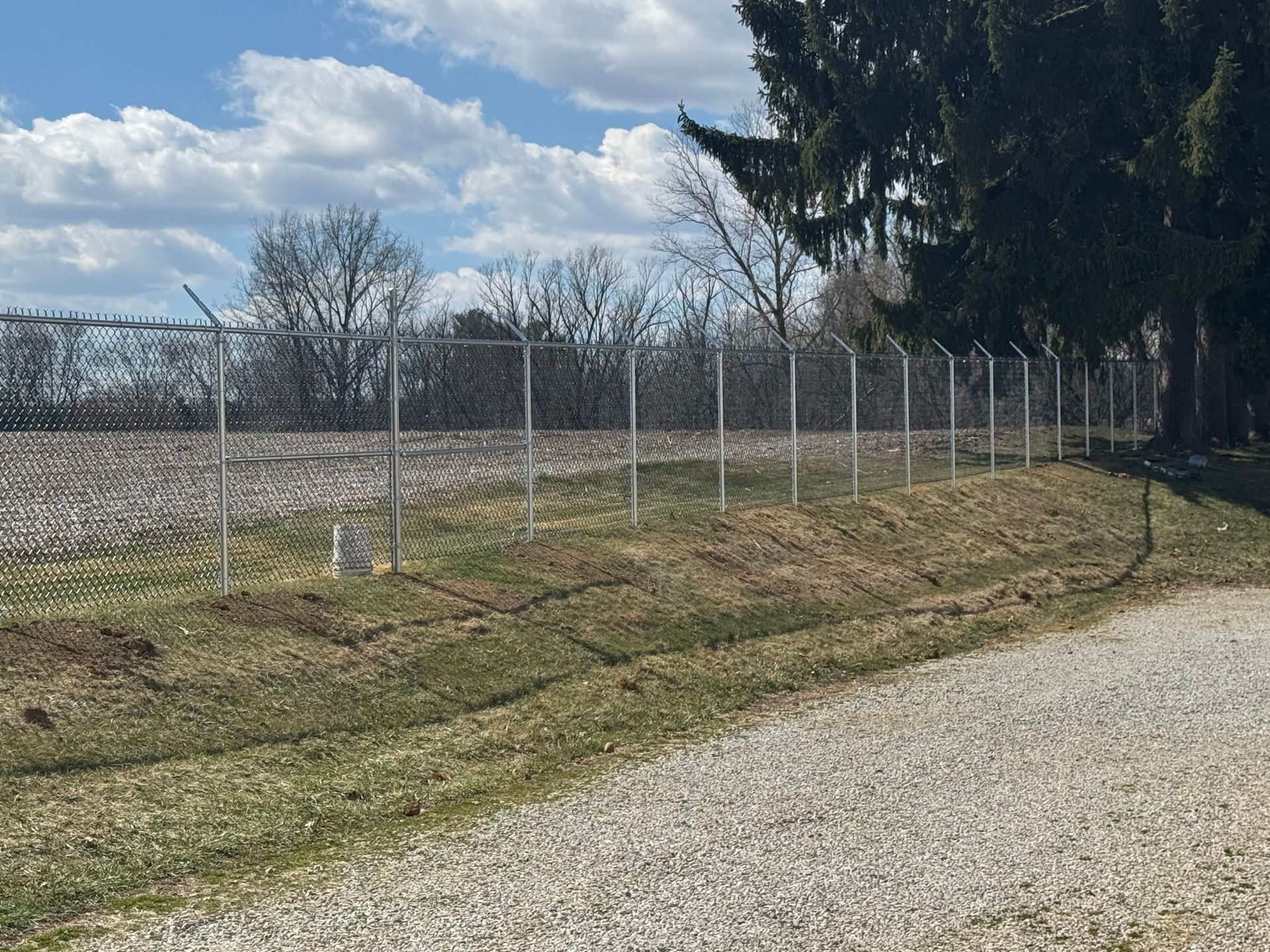 Chain-link fence with barbed wire in field, gravel path in foreground, trees and cloudy sky in background.