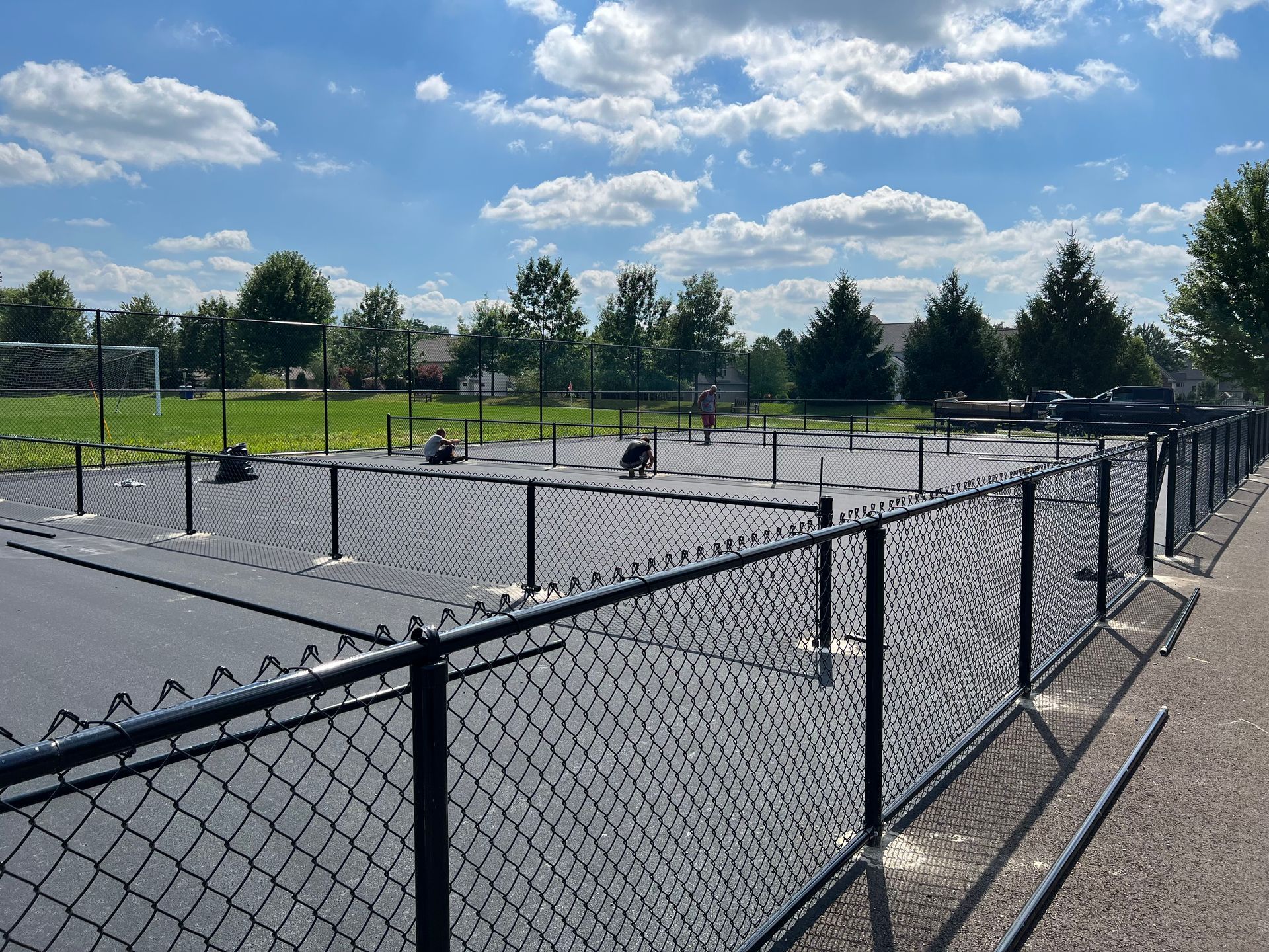 Black chain-link fence surrounds a dark paved area with some equipment. A green field and trees are in the background.