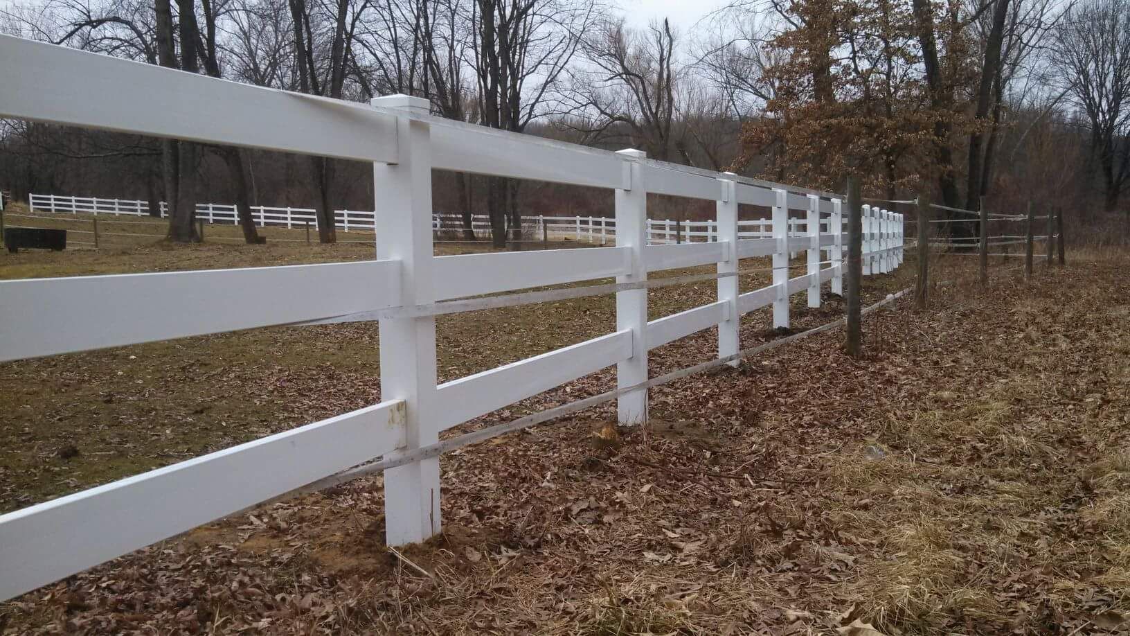 White wooden fence in a field, with brown leaves on the ground and trees in the background.