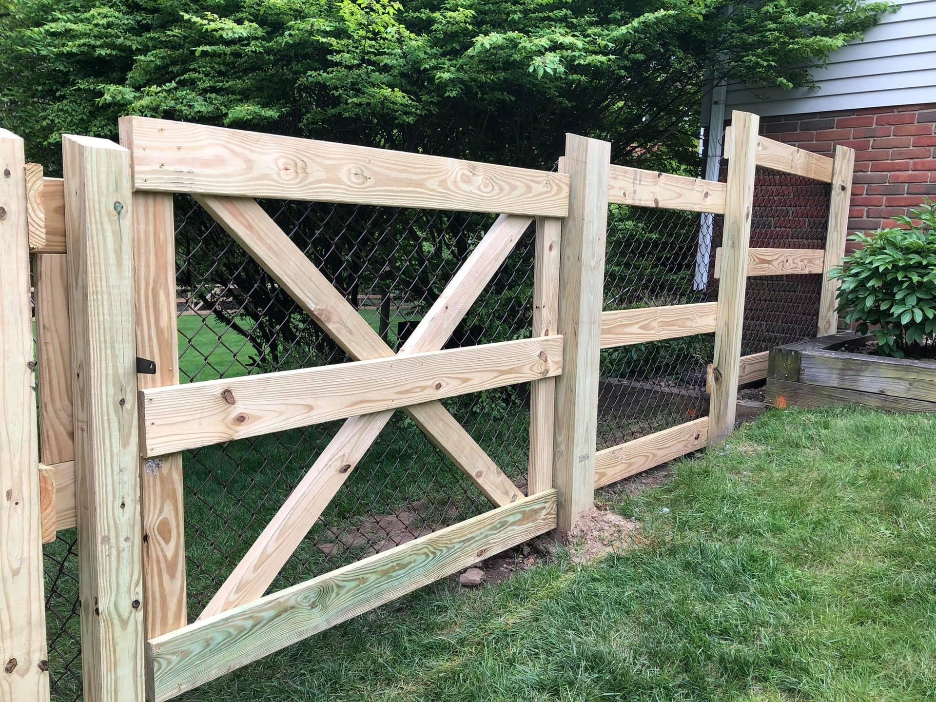 Wooden fence with an X-shaped design and chain link, in a grassy yard next to a brick house.