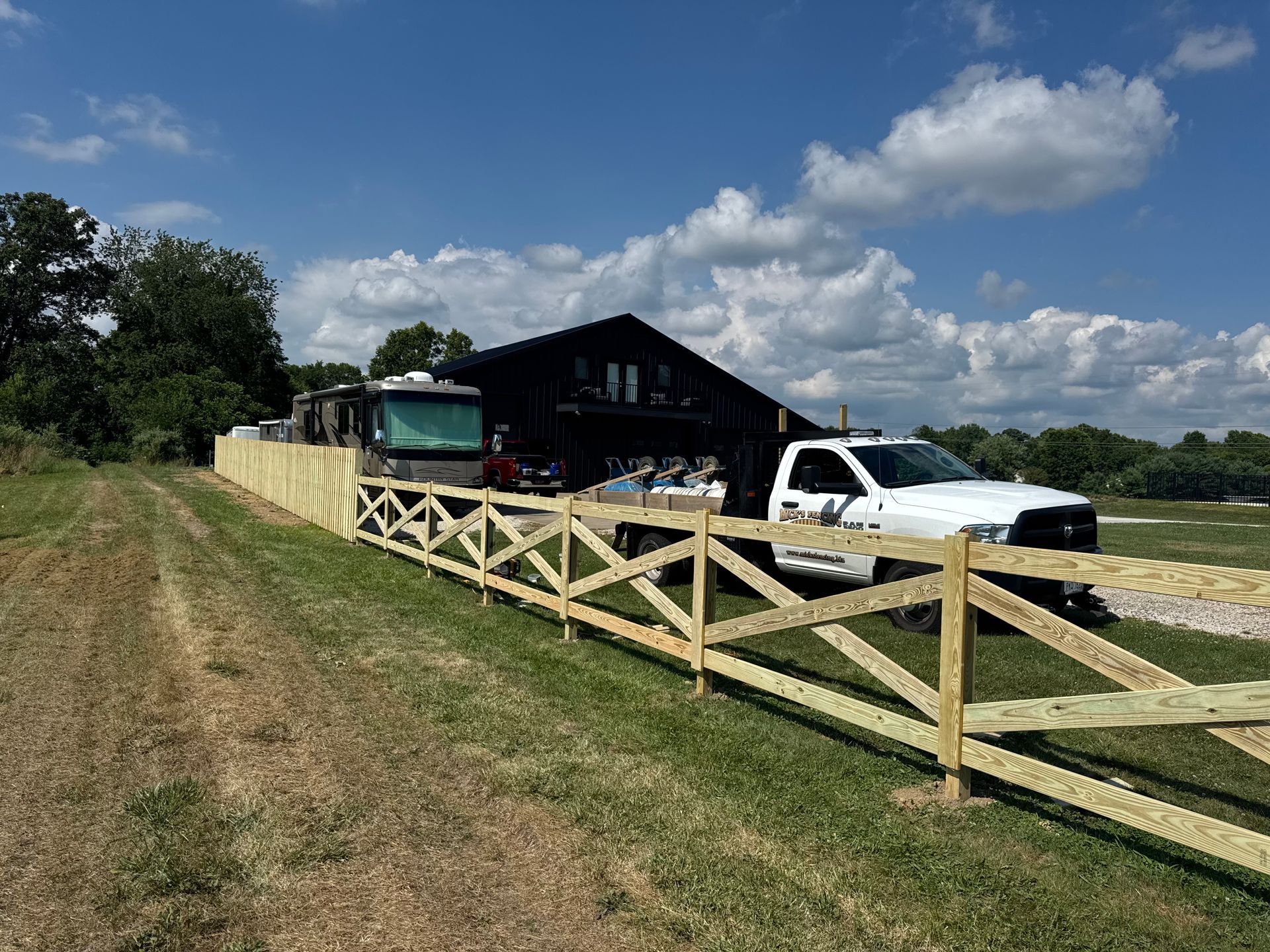 Wooden fence along a dirt path, with a house and white truck in the background under a blue sky.