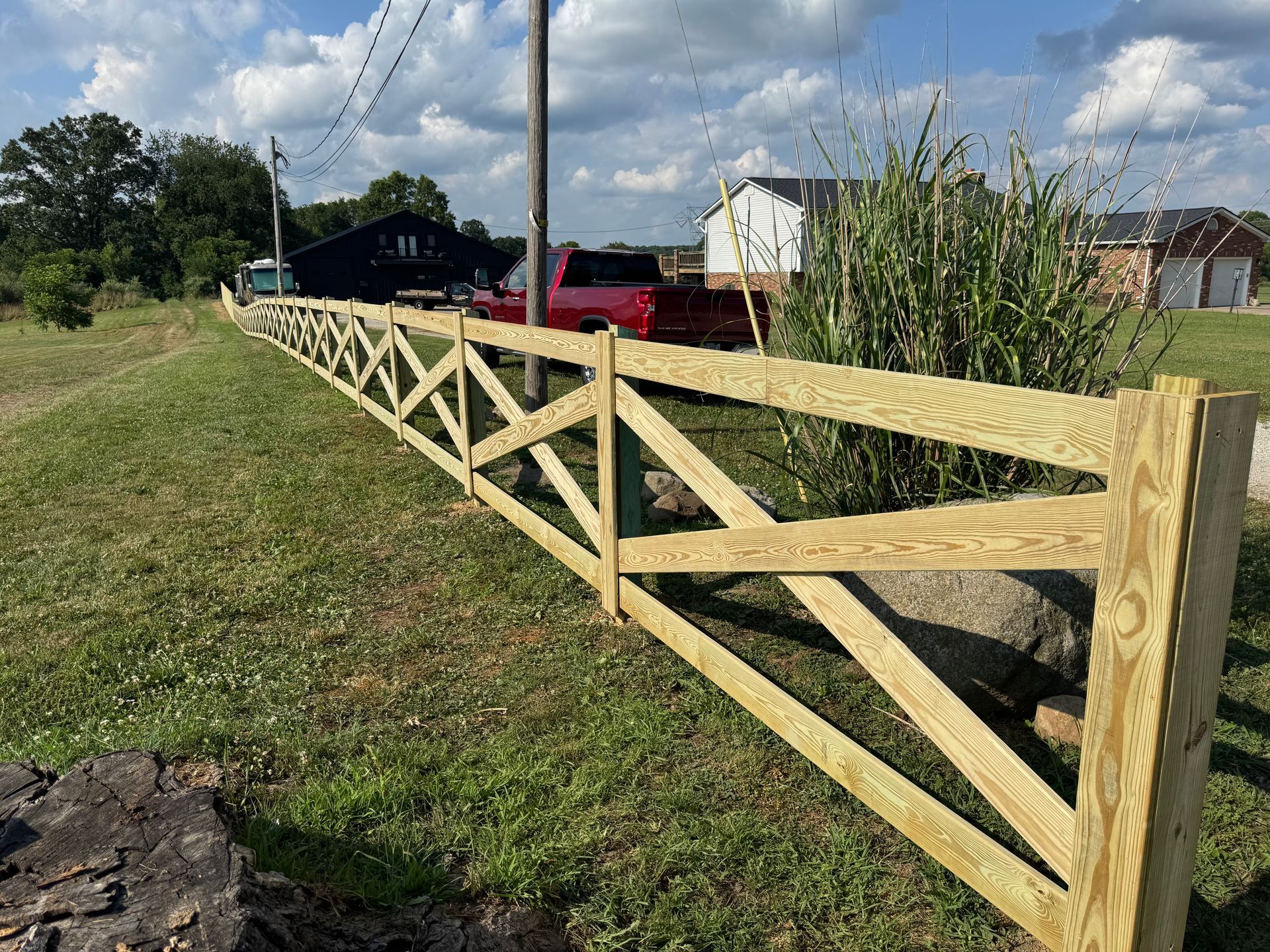 Wooden fence with crisscross design in a grassy field, with houses and vehicles in the background.
