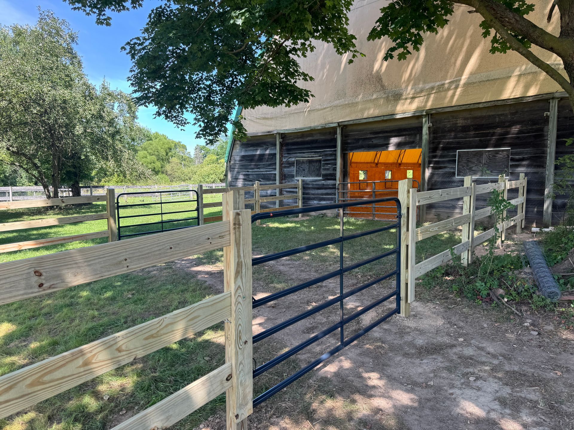 A wooden fence and gate leading to a barn. An orange object is visible. Green grass and trees surround the structure.