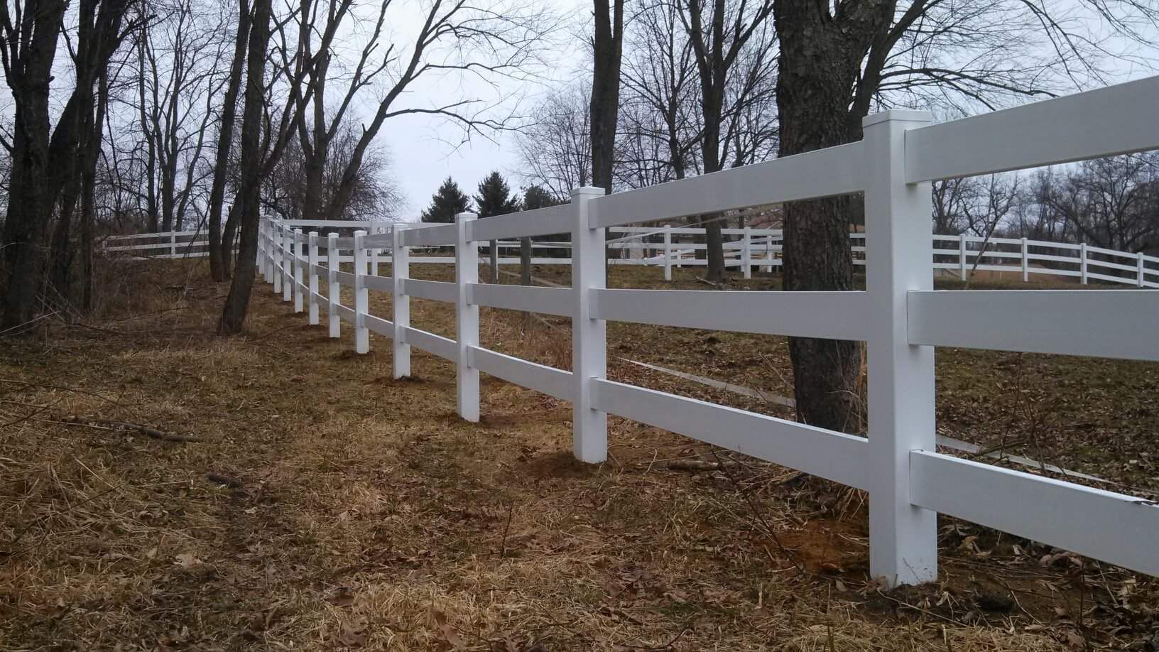 White three-rail fence in a wooded area, with a cloudy sky in the background.