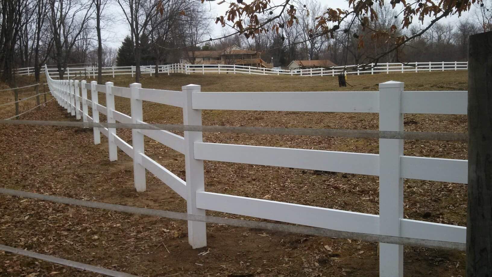 White fence in a field with brown leaves, trees in the background.