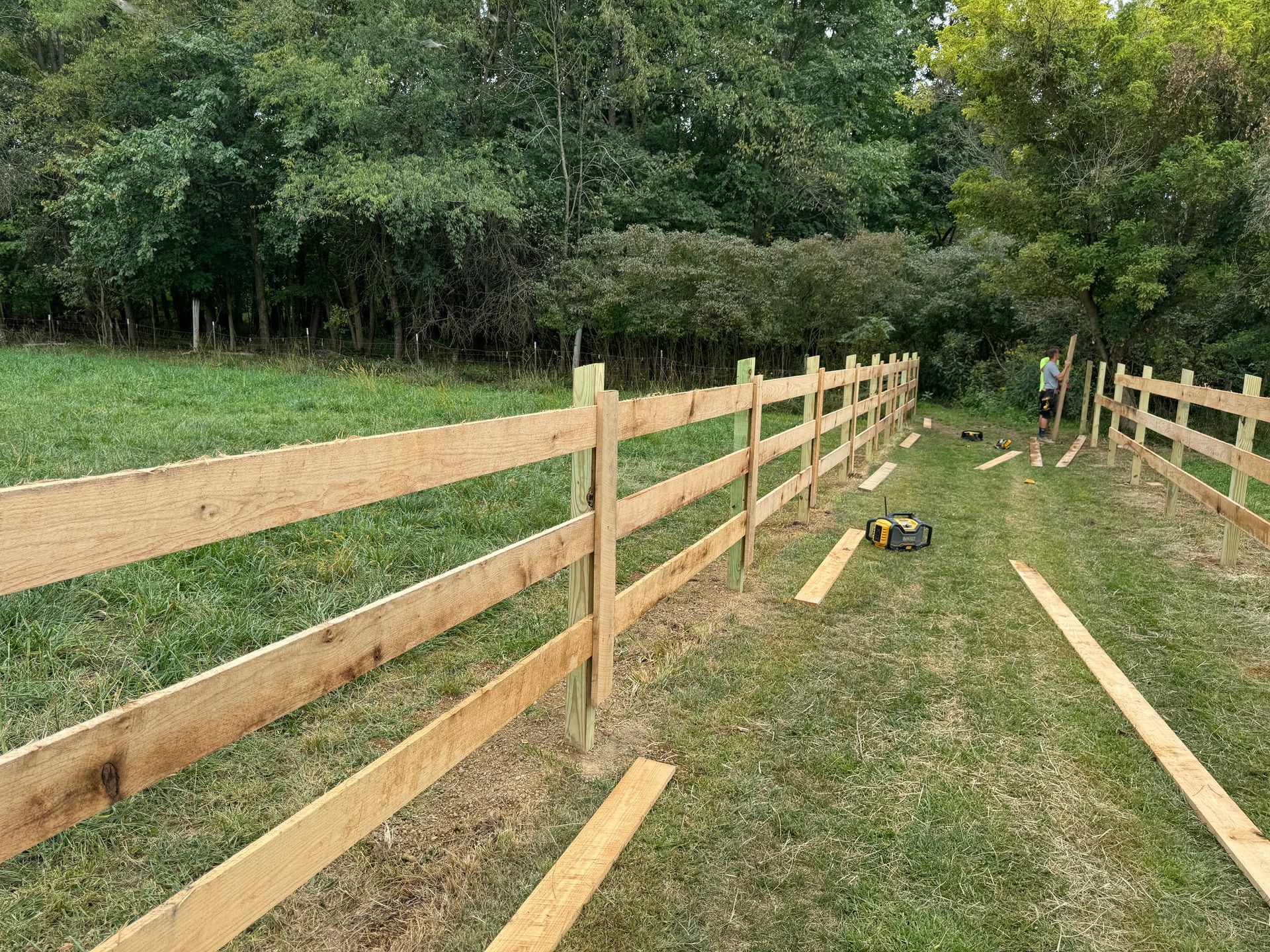 A wooden split-rail fence under construction in a grassy field with trees in the background.