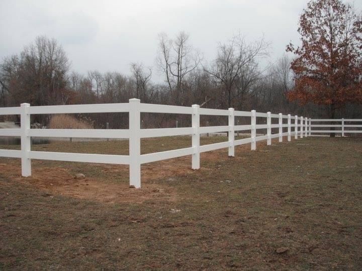 White three-rail fence in a grassy field, with trees in the background under a cloudy sky.