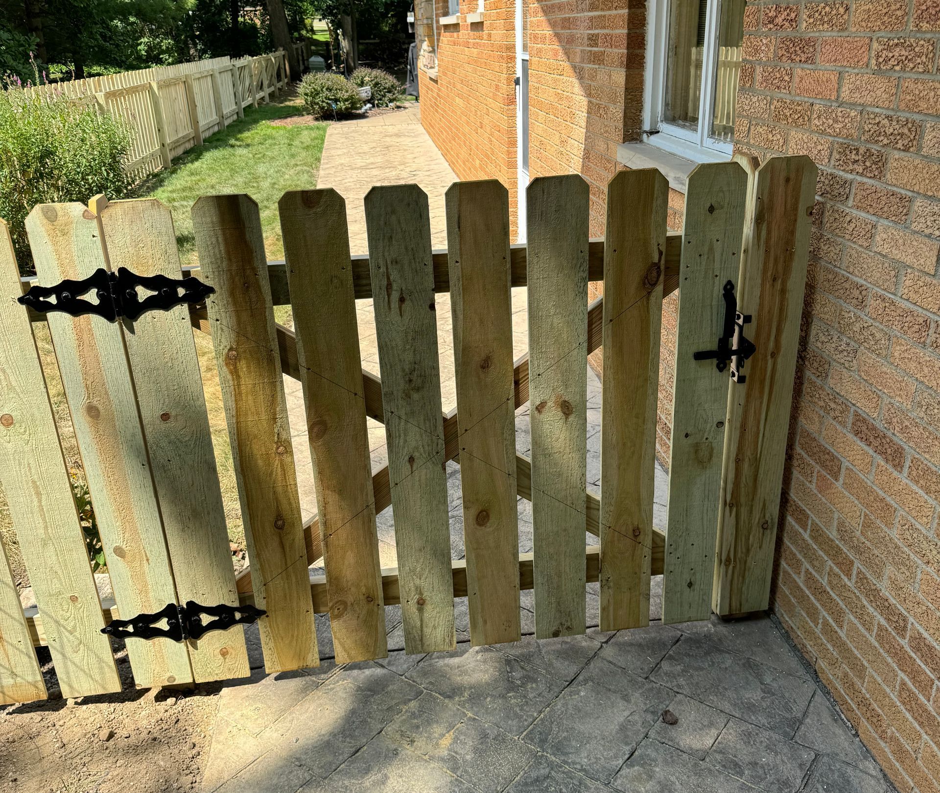 Wooden gate with black hinges and latch, adjacent to brick wall and cement patio.