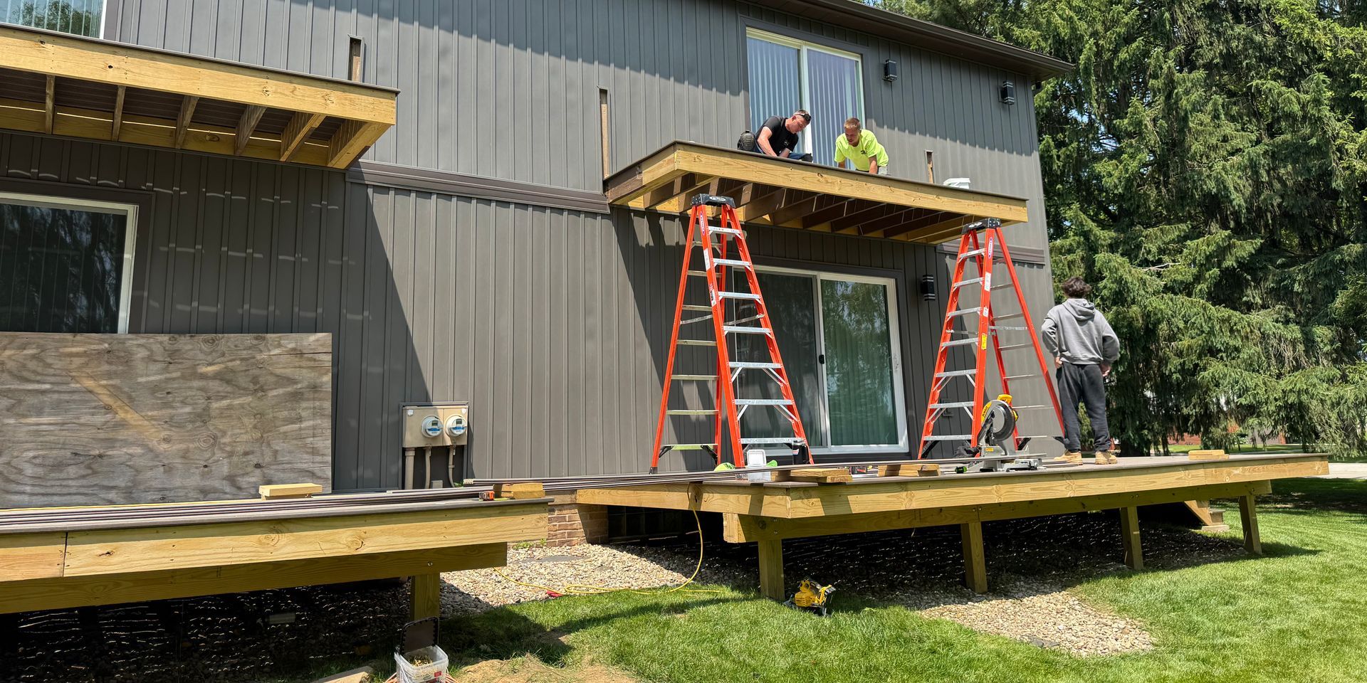 Construction workers build a deck on a house. Red ladders, gray siding, wooden structure, green grass.