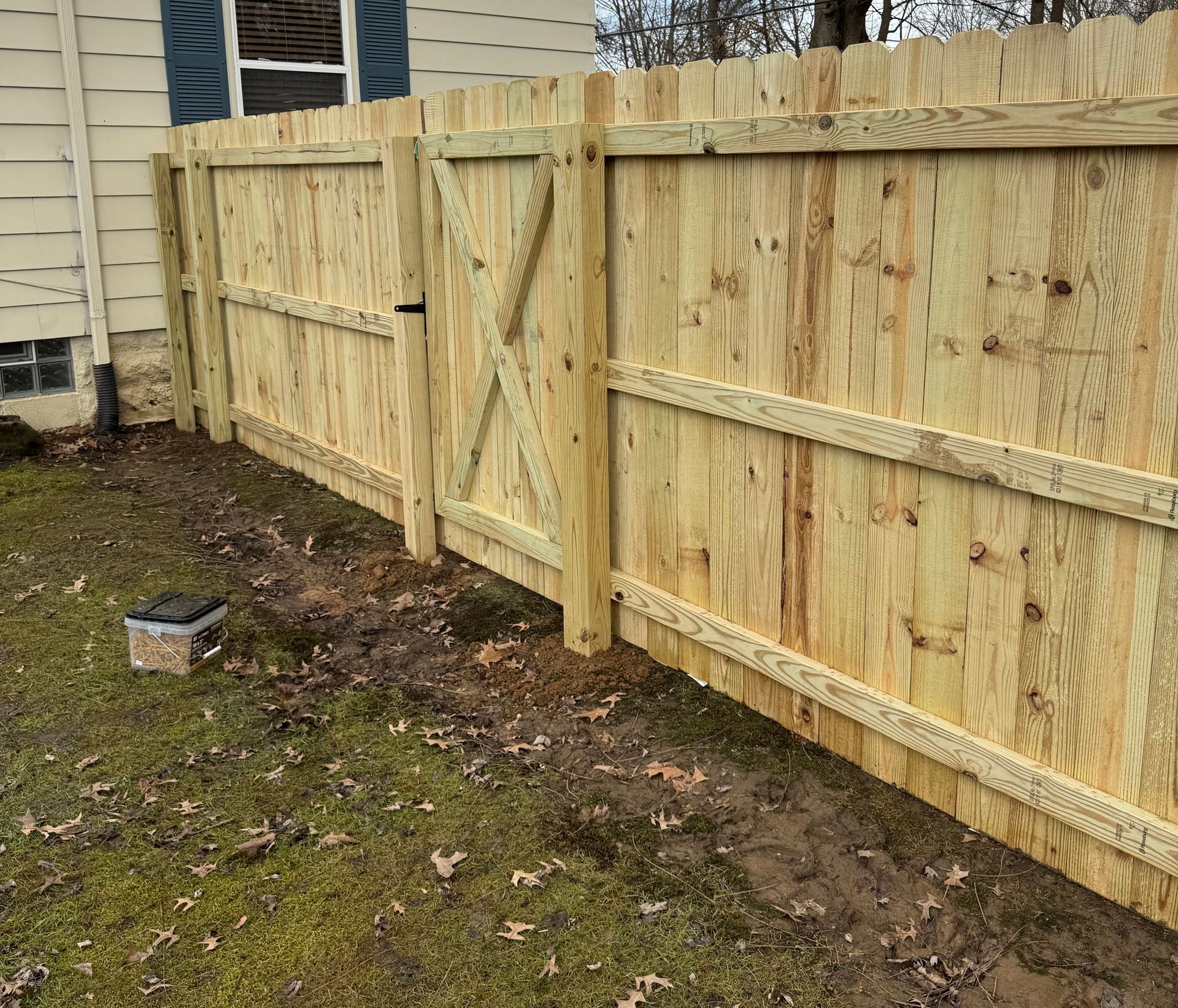 Wooden fence with gate alongside a beige house on a grassy area.