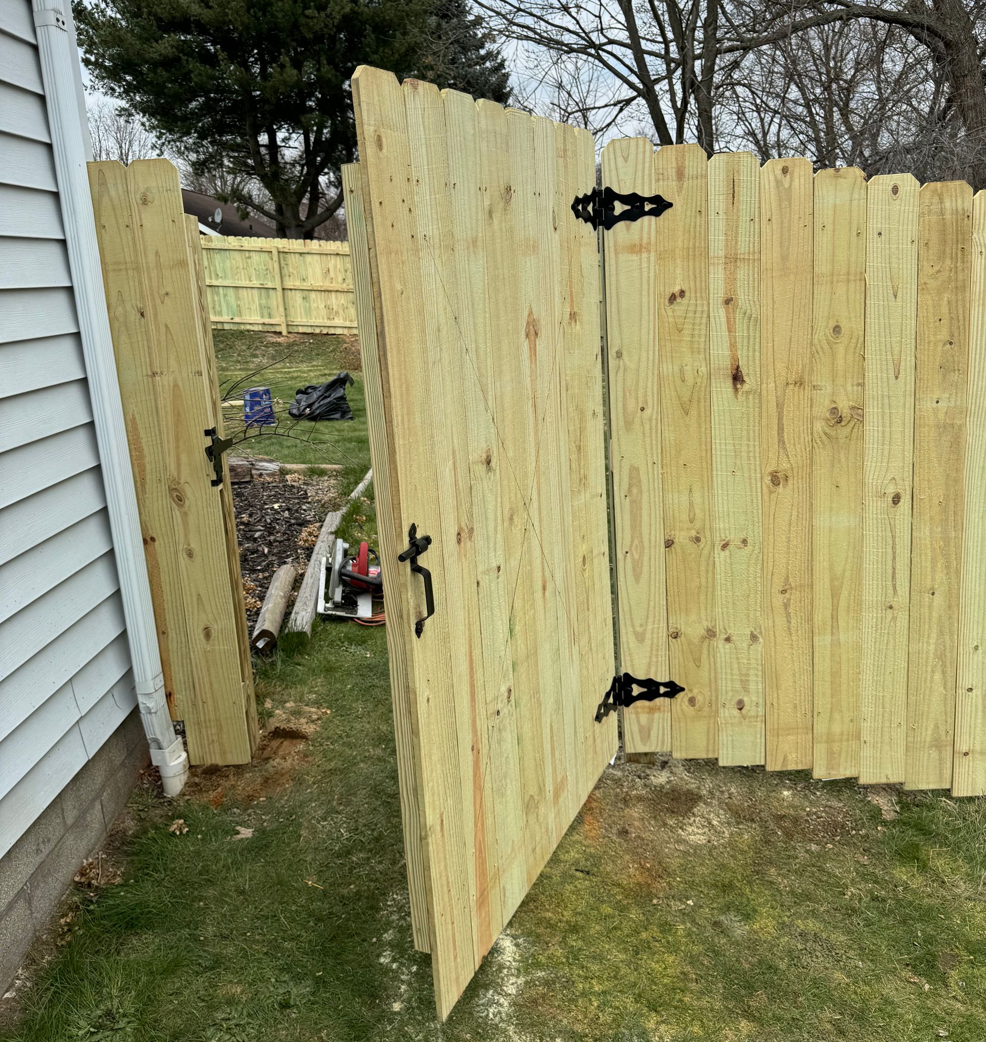 A wooden fence gate, open in a grassy yard, with black hinges and latch.