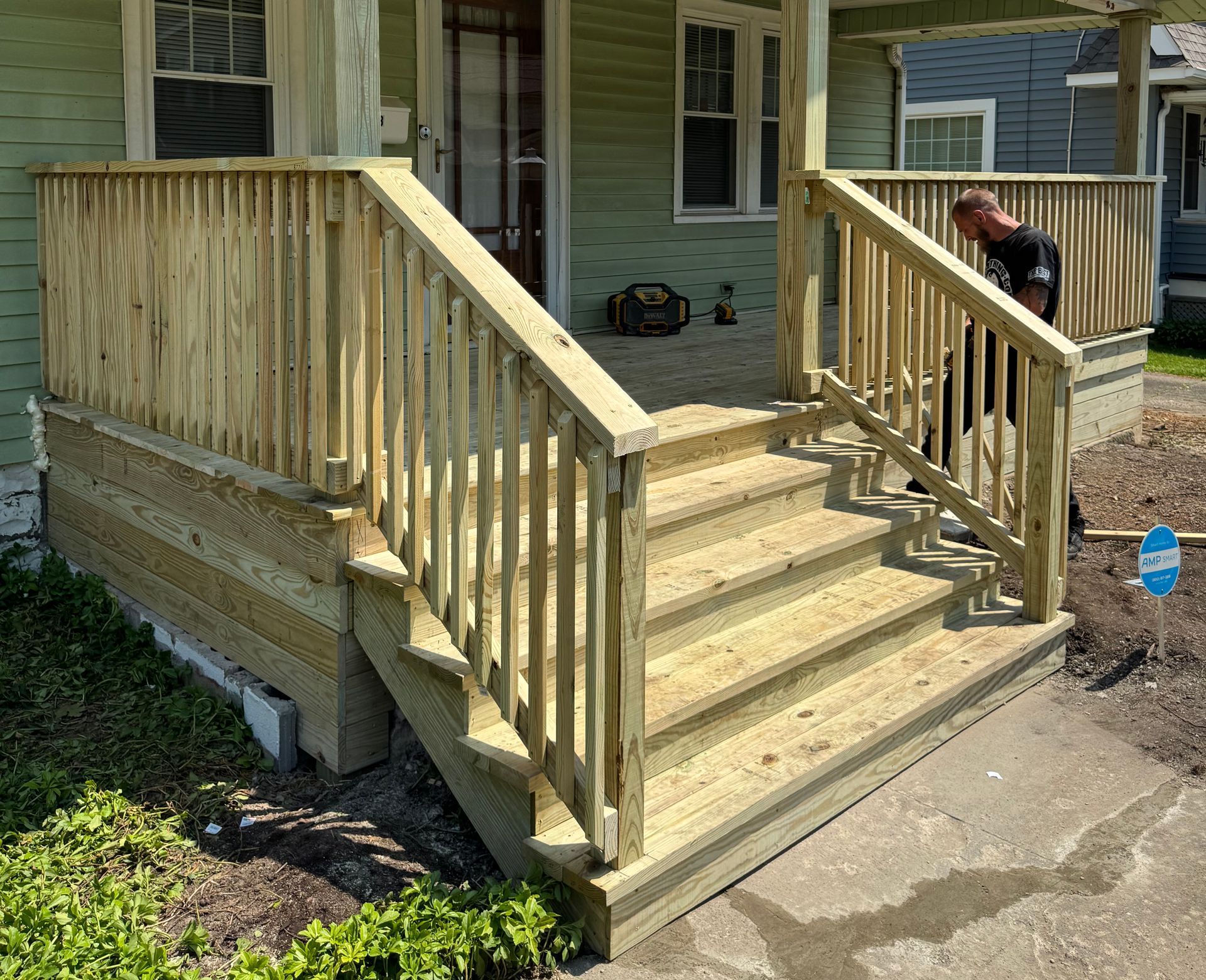 Wooden porch with steps, railings, and a man. Green house exterior and cement walkway.