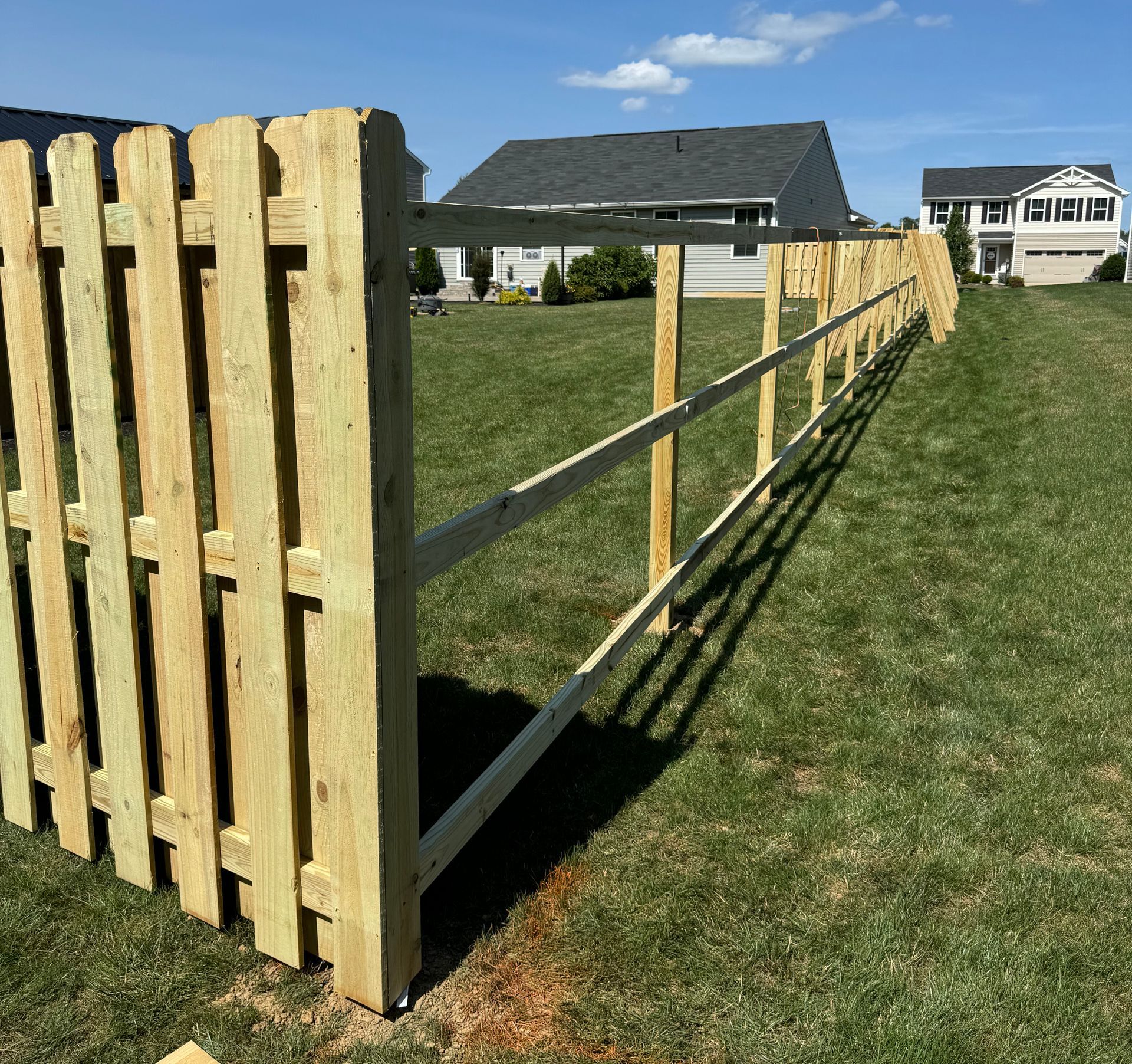 Wooden fence being constructed in a grassy yard on a sunny day, with houses in the background.