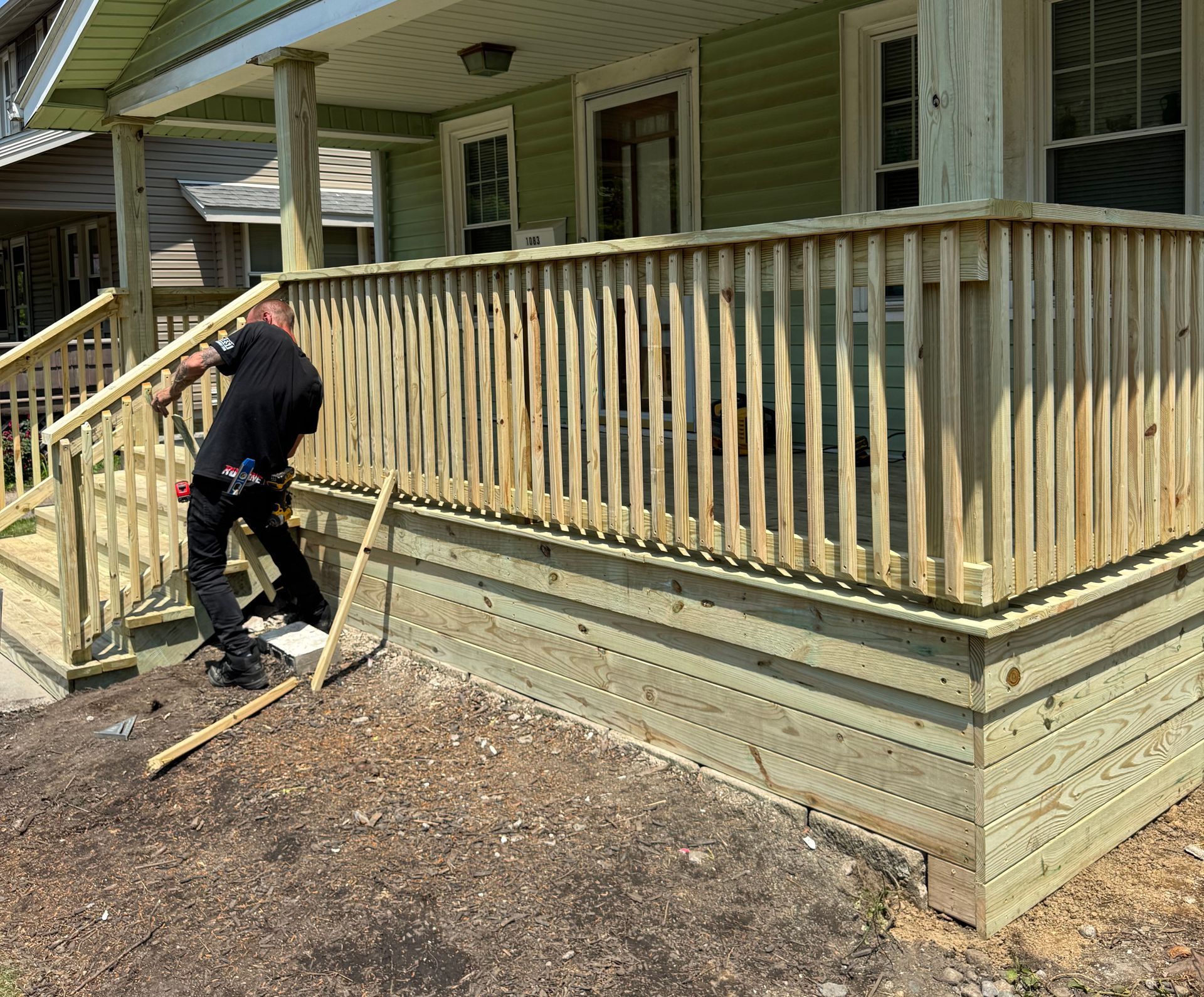 Man working on a new wooden porch with railing. Green house in the background. Sunny day.