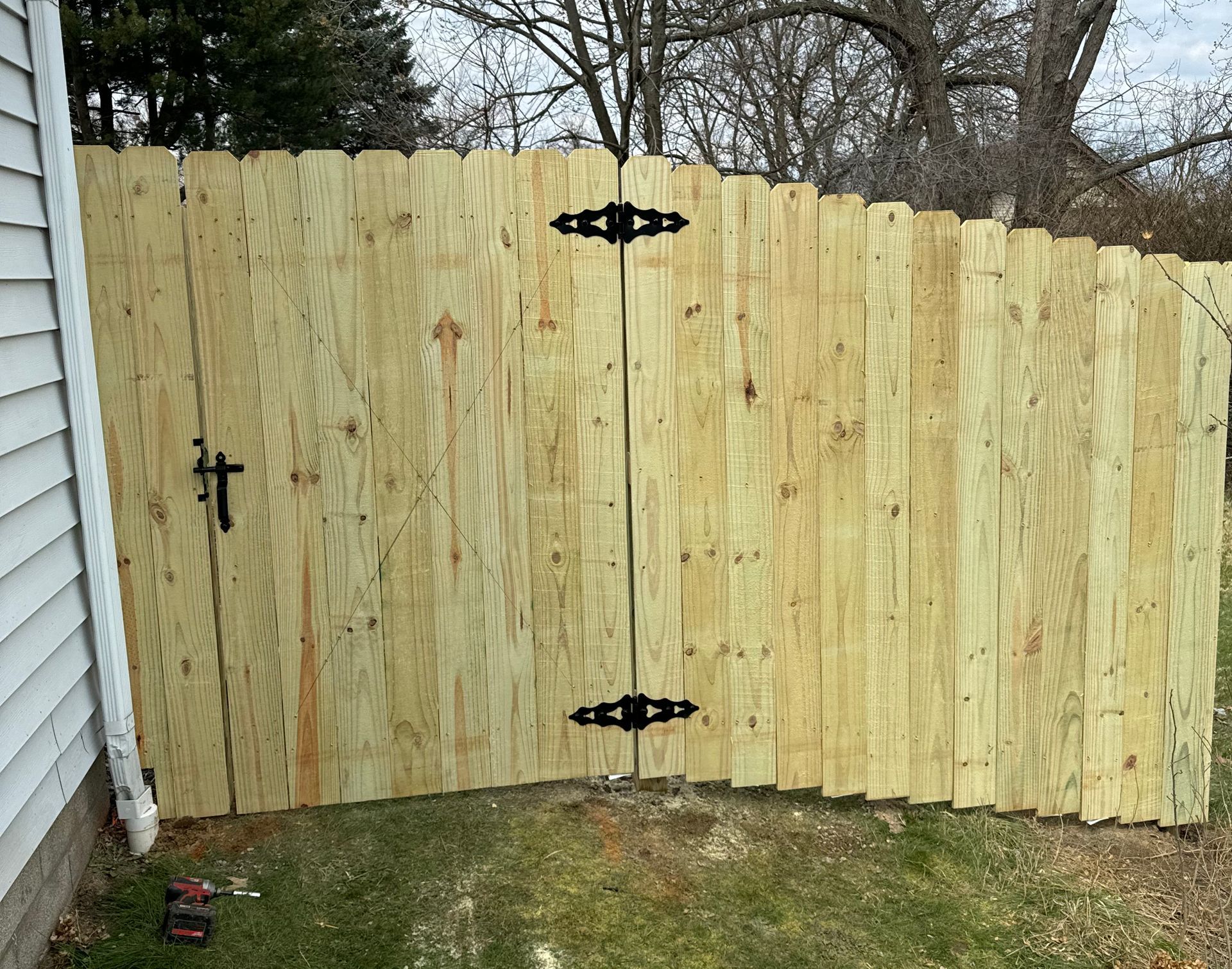 Wooden fence gate with black hardware, set in a yard next to a white house.