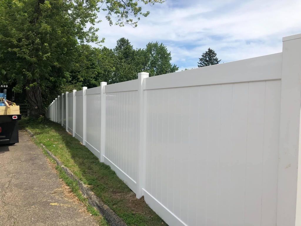 White vinyl fence lining a street, with green trees and cloudy sky in the background.