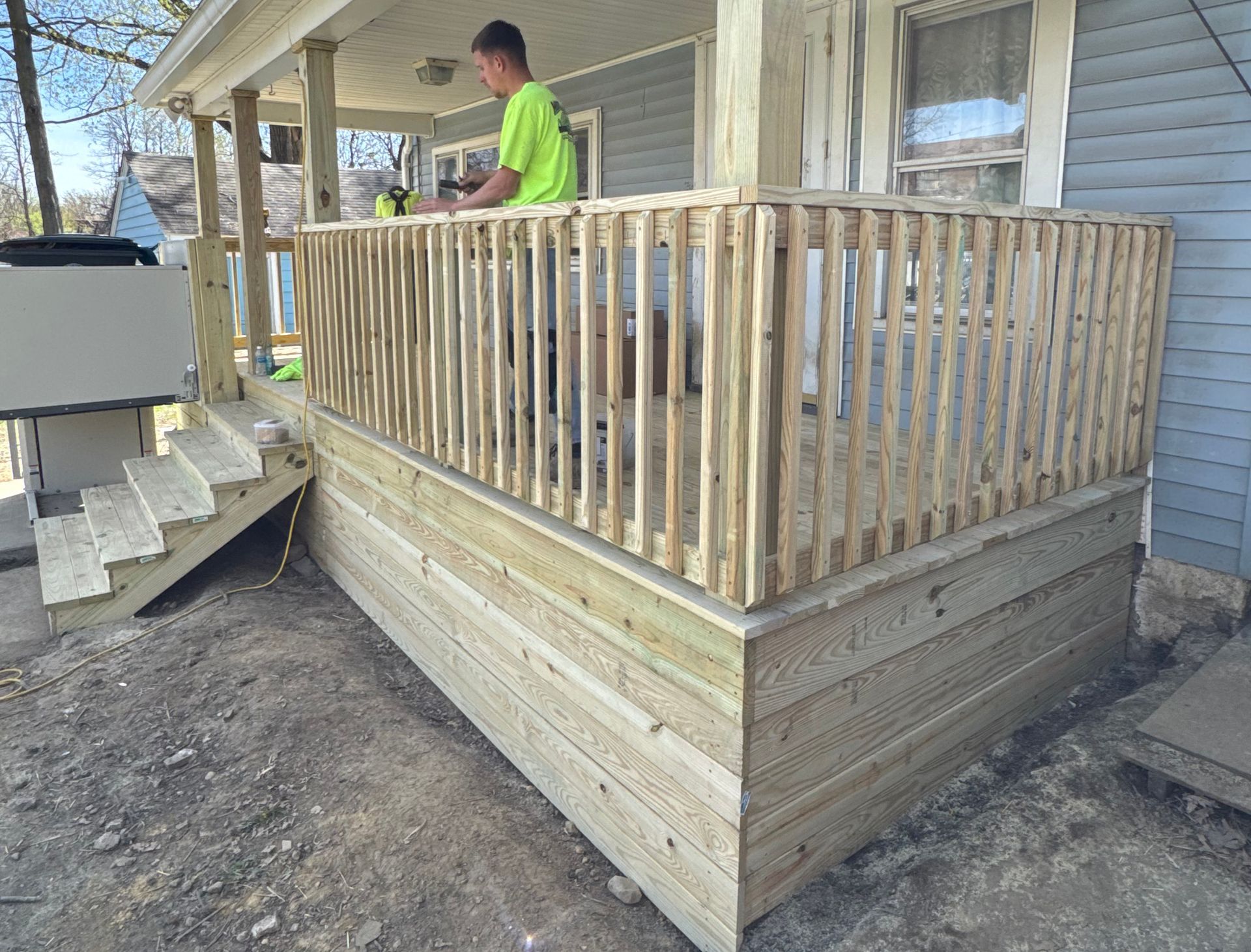 A person on a wooden deck, building a railing. The deck is attached to a blue house.