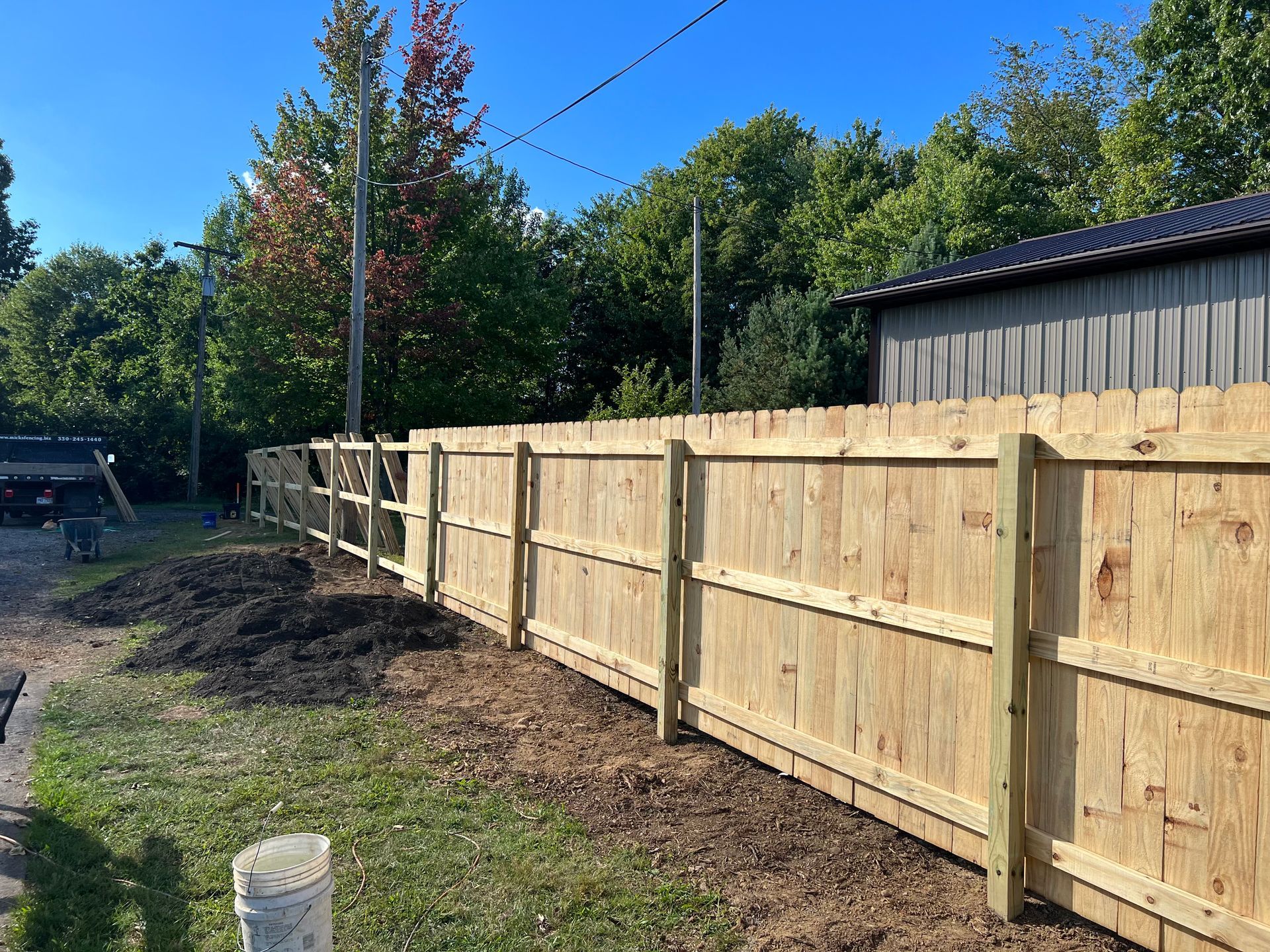 A wooden fence, newly built, lines the edge of a yard, with a building and trees in the background under a blue sky.
