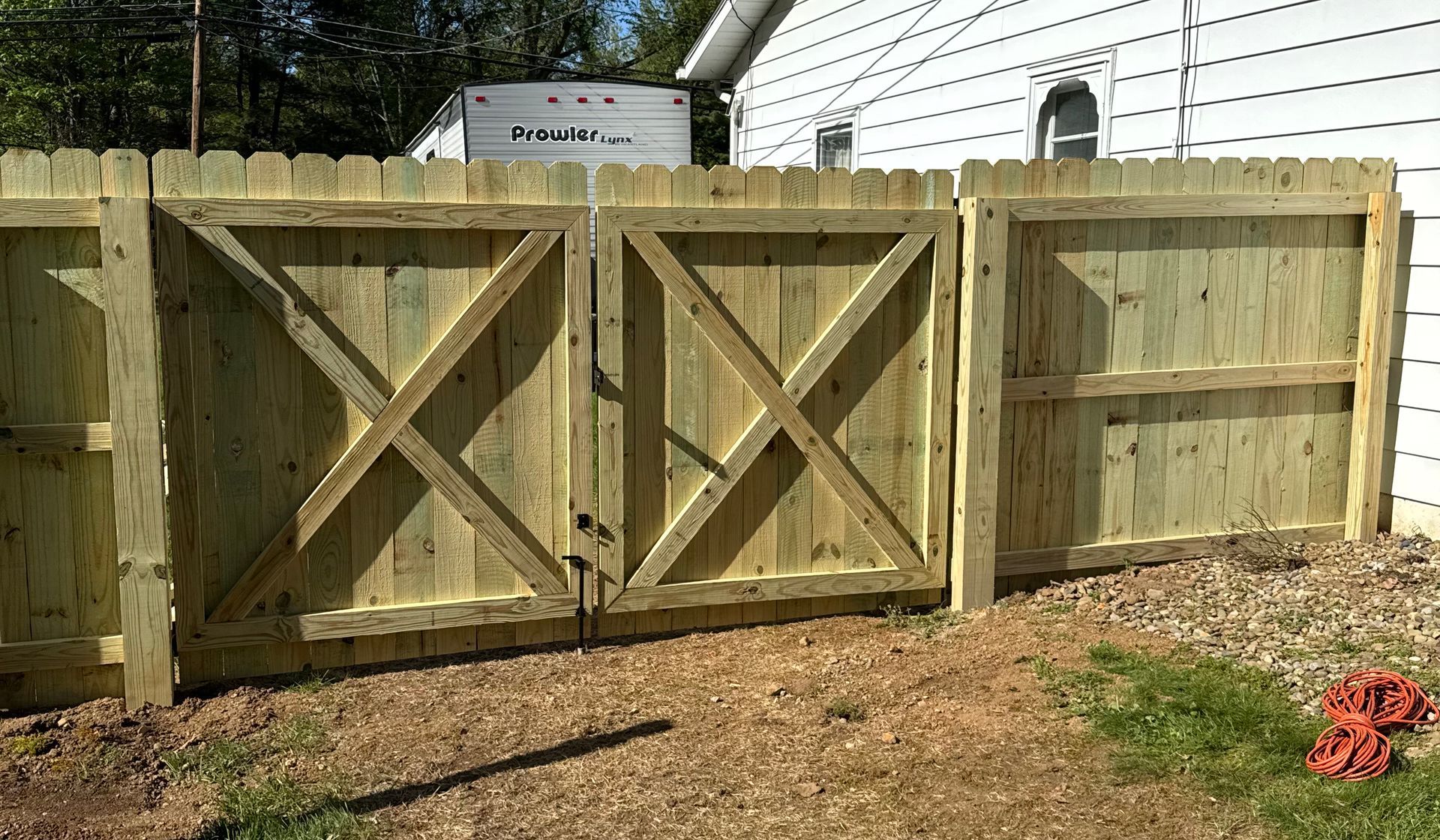 Wooden fence with double gate and an X-shaped design; behind it is a white house and a camper.