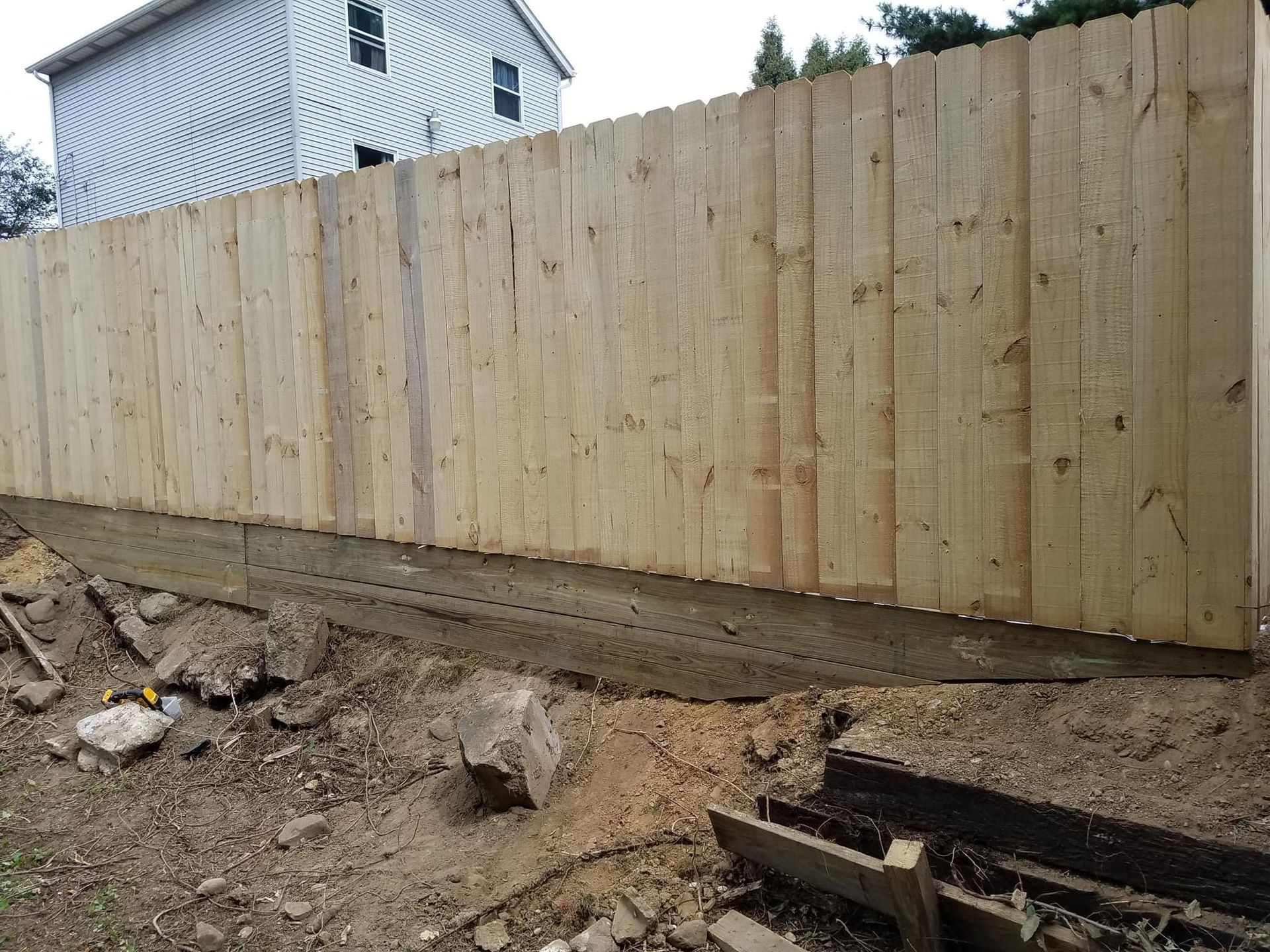 Wooden fence along a yard with exposed soil, and a house in the background.