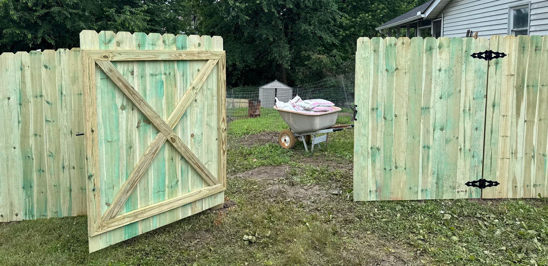 Green wooden fence with gate open. A wheelbarrow is inside.