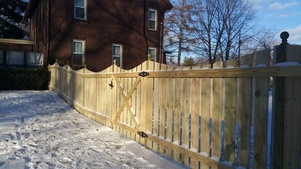 Wooden fence with gate in snowy yard, with a brown brick house in the background.