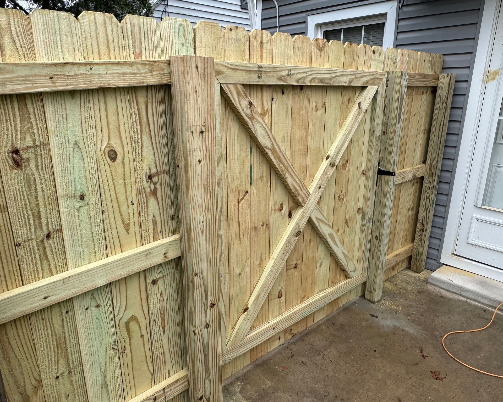 Wooden gate in a wooden fence, next to a light blue house with a white door.