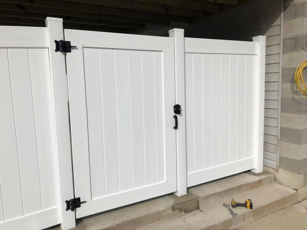 White vinyl fence gates with black hinges and handle, set against a concrete wall.