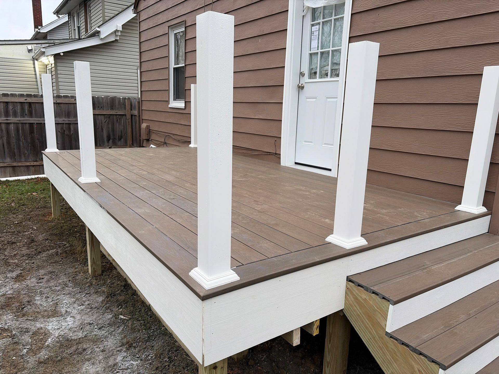 Brown composite deck with white pillars and stairs against a brown house, cloudy sky